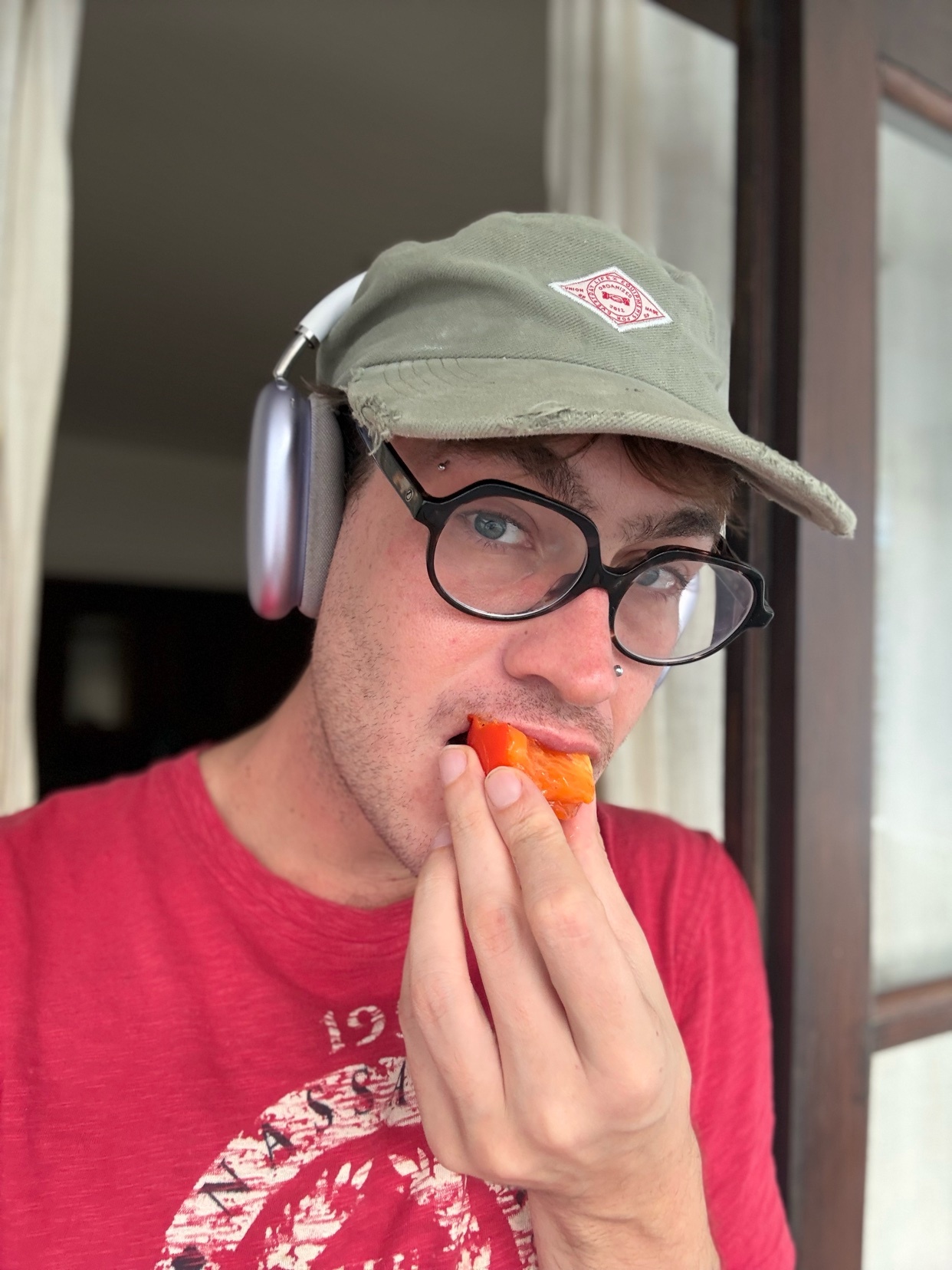 A man wearing a baseball cap and glasses eats a piece of fruit while wearing headphones.