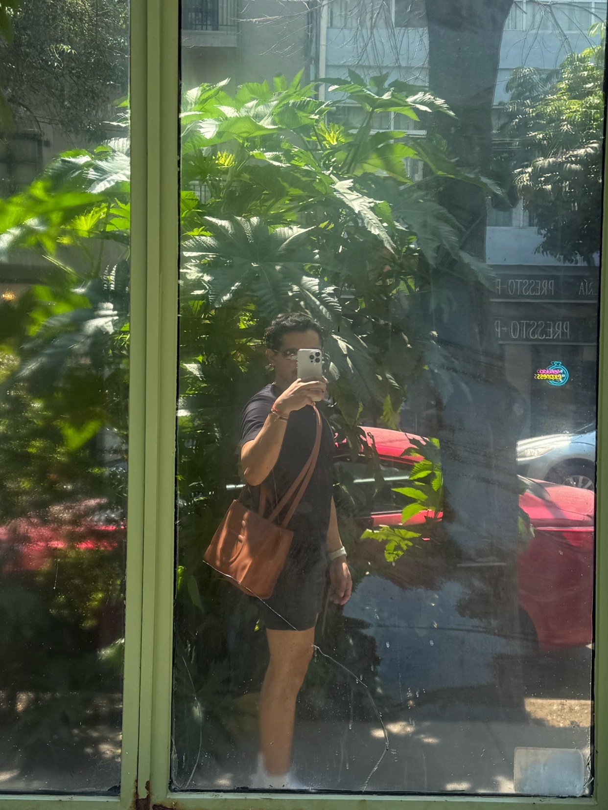 A man takes a selfie in a mirror, wearing a t-shirt and shorts, with a bag slung over his shoulder. The reflection shows a street scene with greenery and a red car.