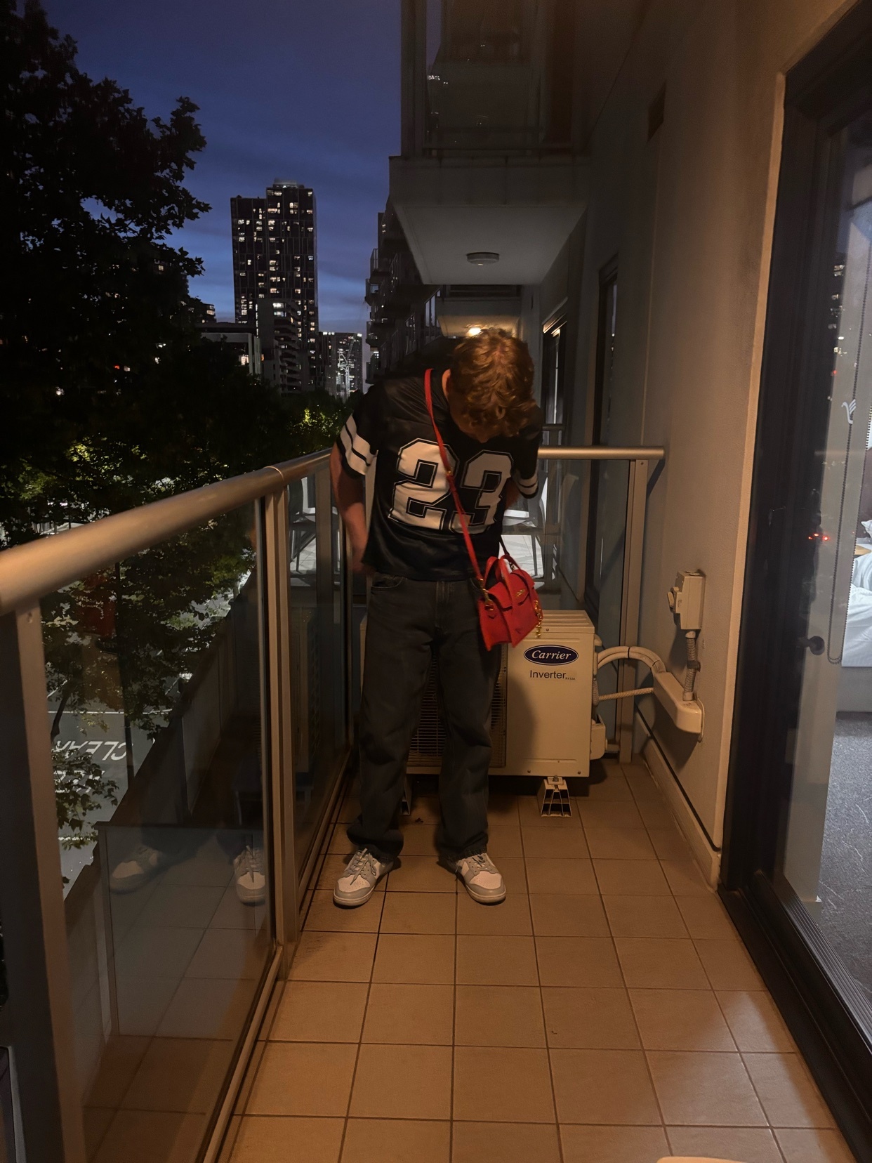 A man stands on a balcony at night, wearing a football jersey and jeans. The cityscape is visible in the background.