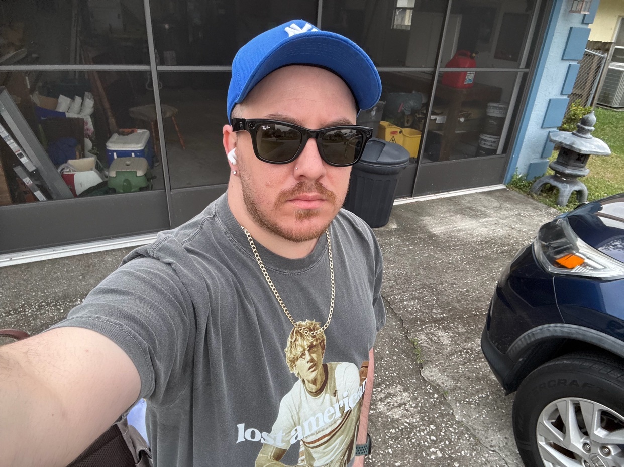 A man wearing a blue baseball cap, sunglasses, and a graphic t-shirt takes a selfie in front of a garage.
