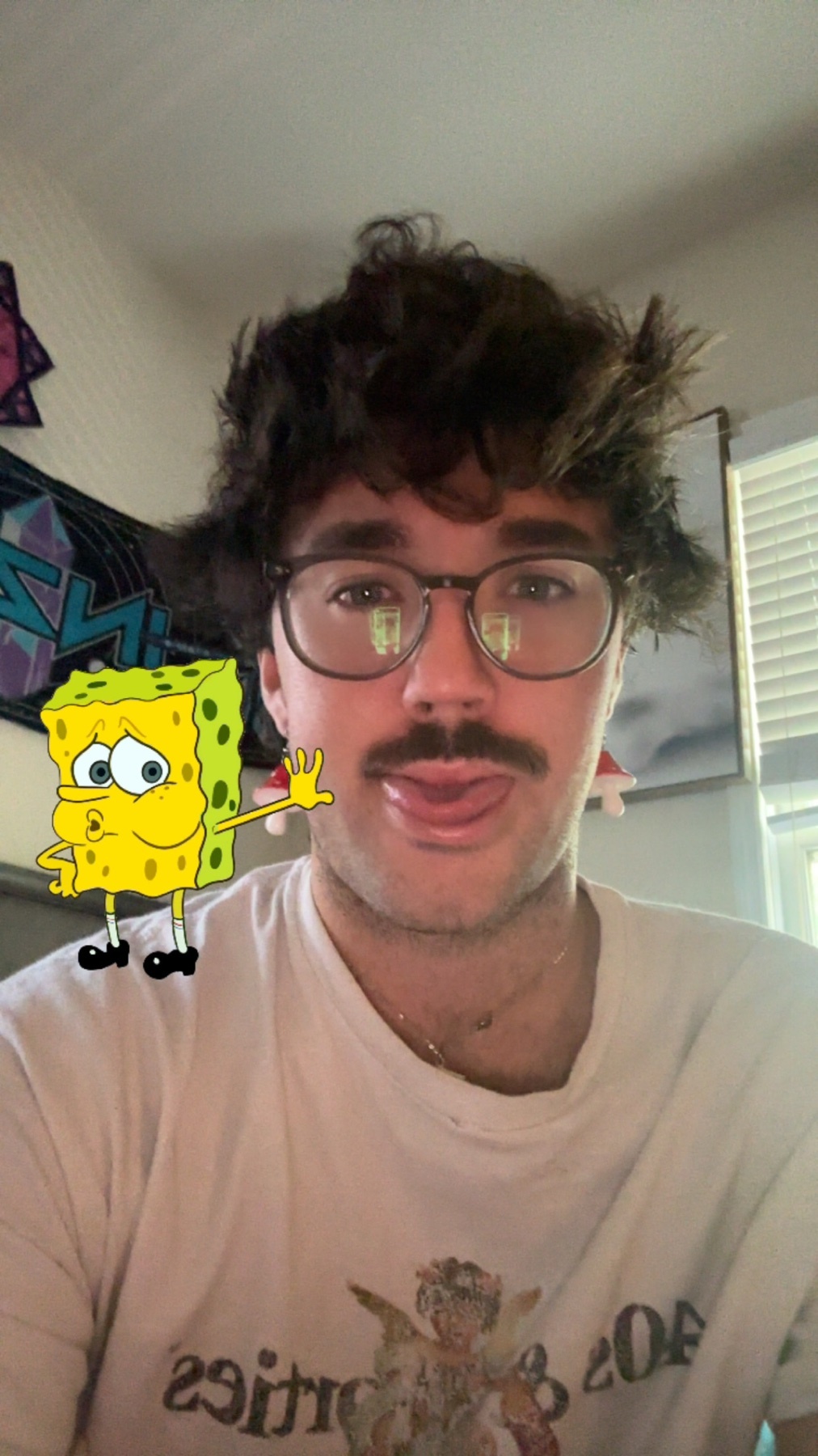 A man with glasses and a mustache sticks his tongue out while posing for a selfie with Spongebob Squarepants.
