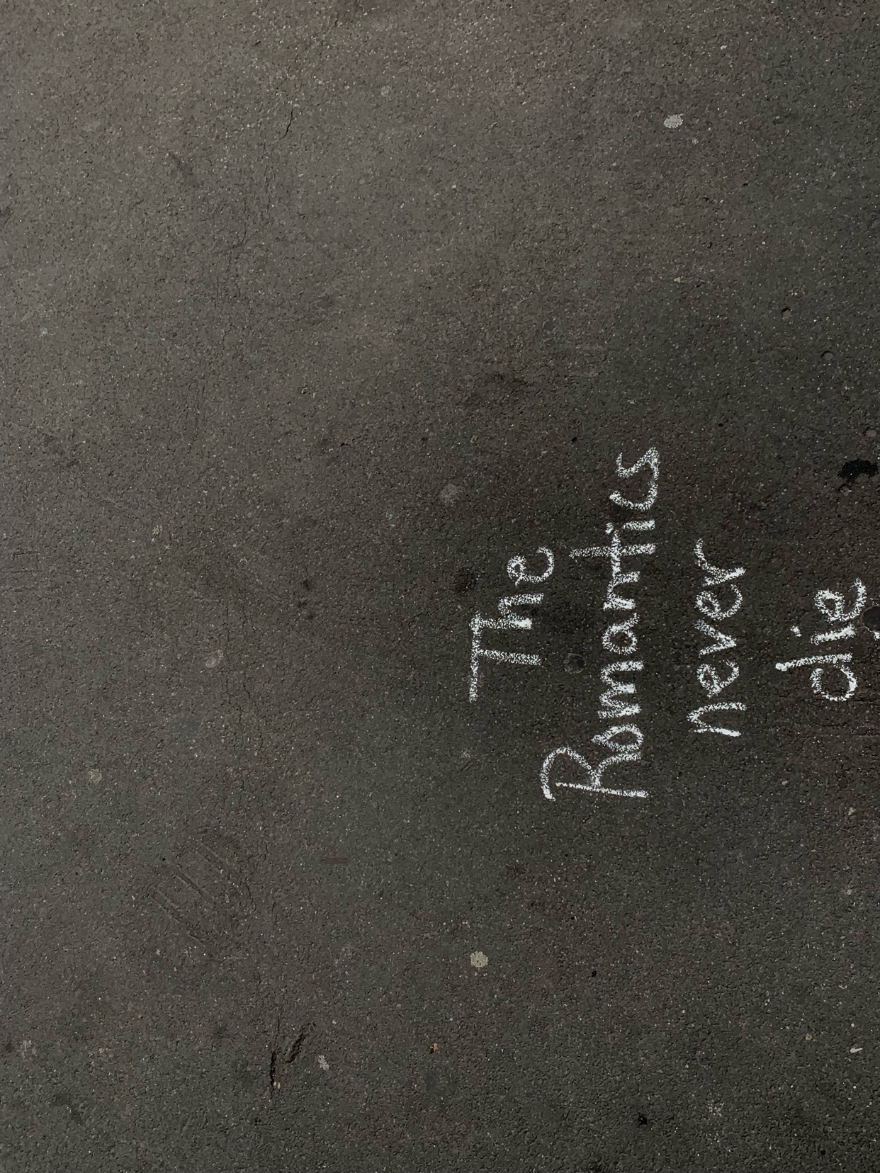 A close-up shot of chalk writing on a dark surface. The text reads "The Romantics never die."