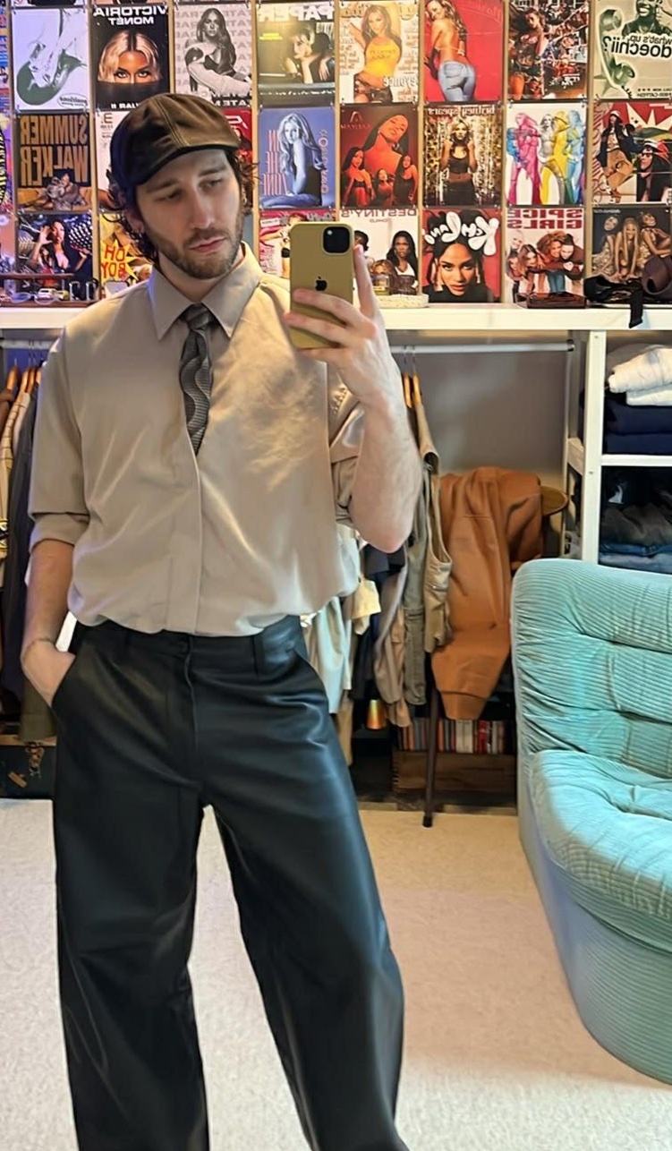 A man in a tan button-down shirt, tie, and black leather pants takes a selfie in front of a wall covered in vintage posters. He is wearing a flat cap and has his hand in his pocket.