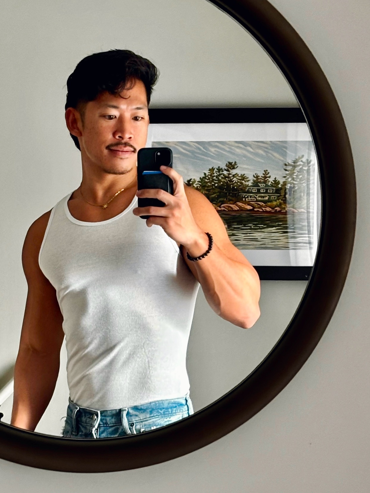 A man takes a mirror selfie, showcasing his physique. He is shirtless, wearing a white tank top and jeans.