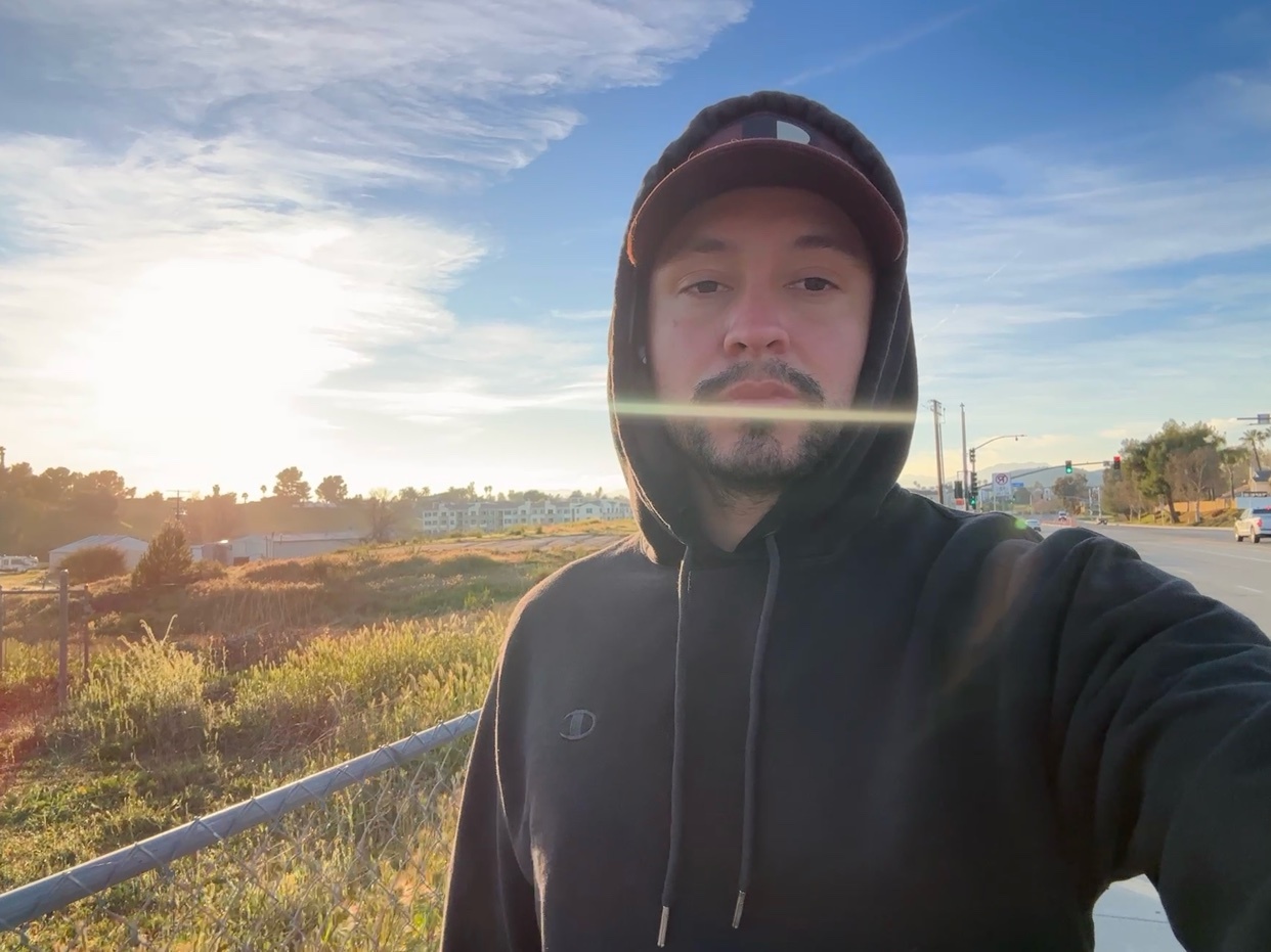 A man in a black hoodie and a baseball cap takes a selfie outdoors during the day. The sun is shining brightly in the background, casting a warm glow.