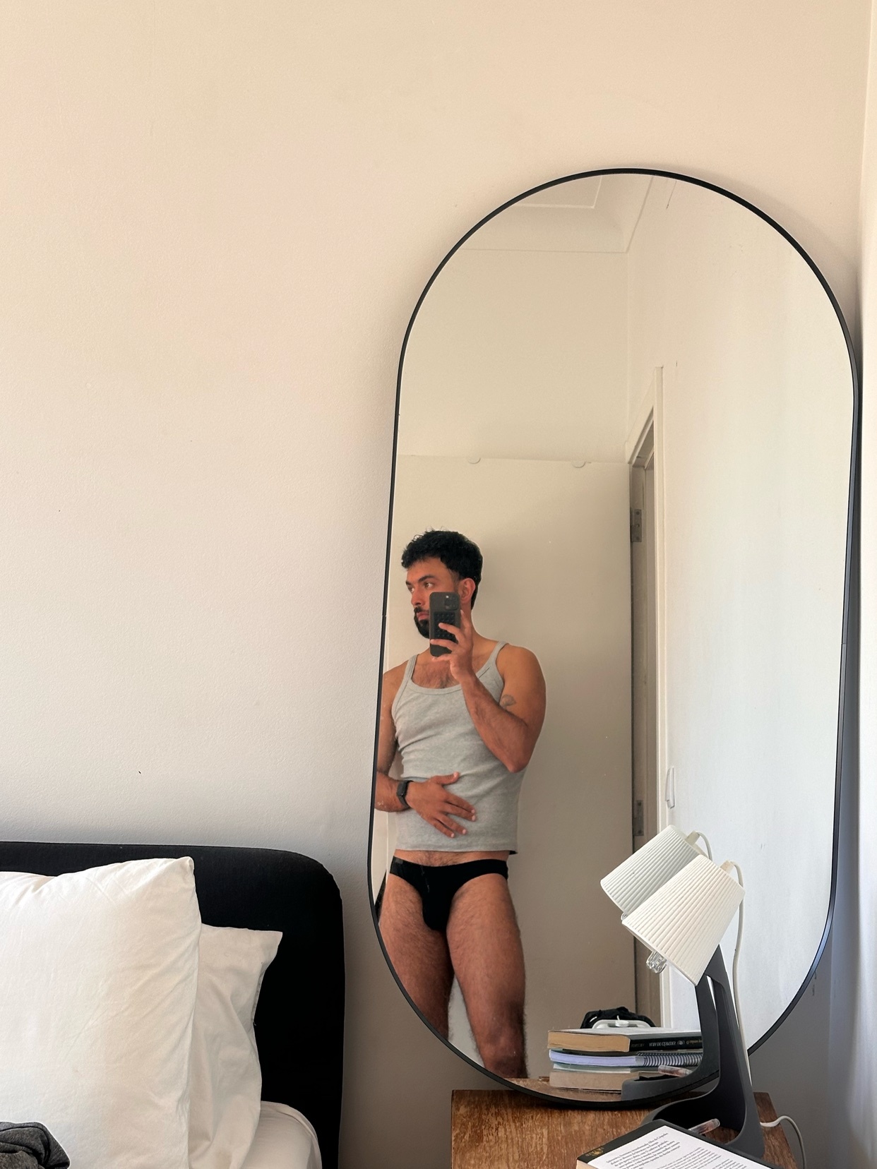 A man in a tank top and underwear takes a mirror selfie in a bedroom.