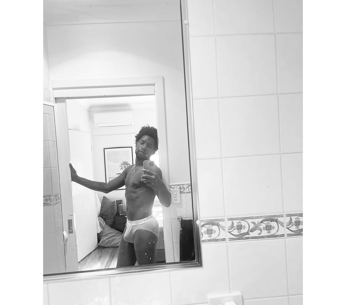 A black and white mirror selfie of a man in underwear. He is standing in a bathroom and appears to be flexing.