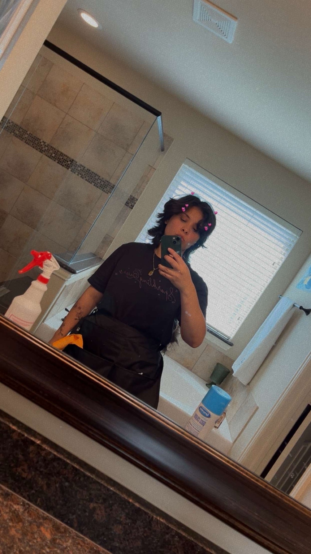 A woman takes a selfie in a bathroom mirror, holding a cleaning spray bottle and wearing work clothes. The bathroom features a tiled shower and a bathtub.