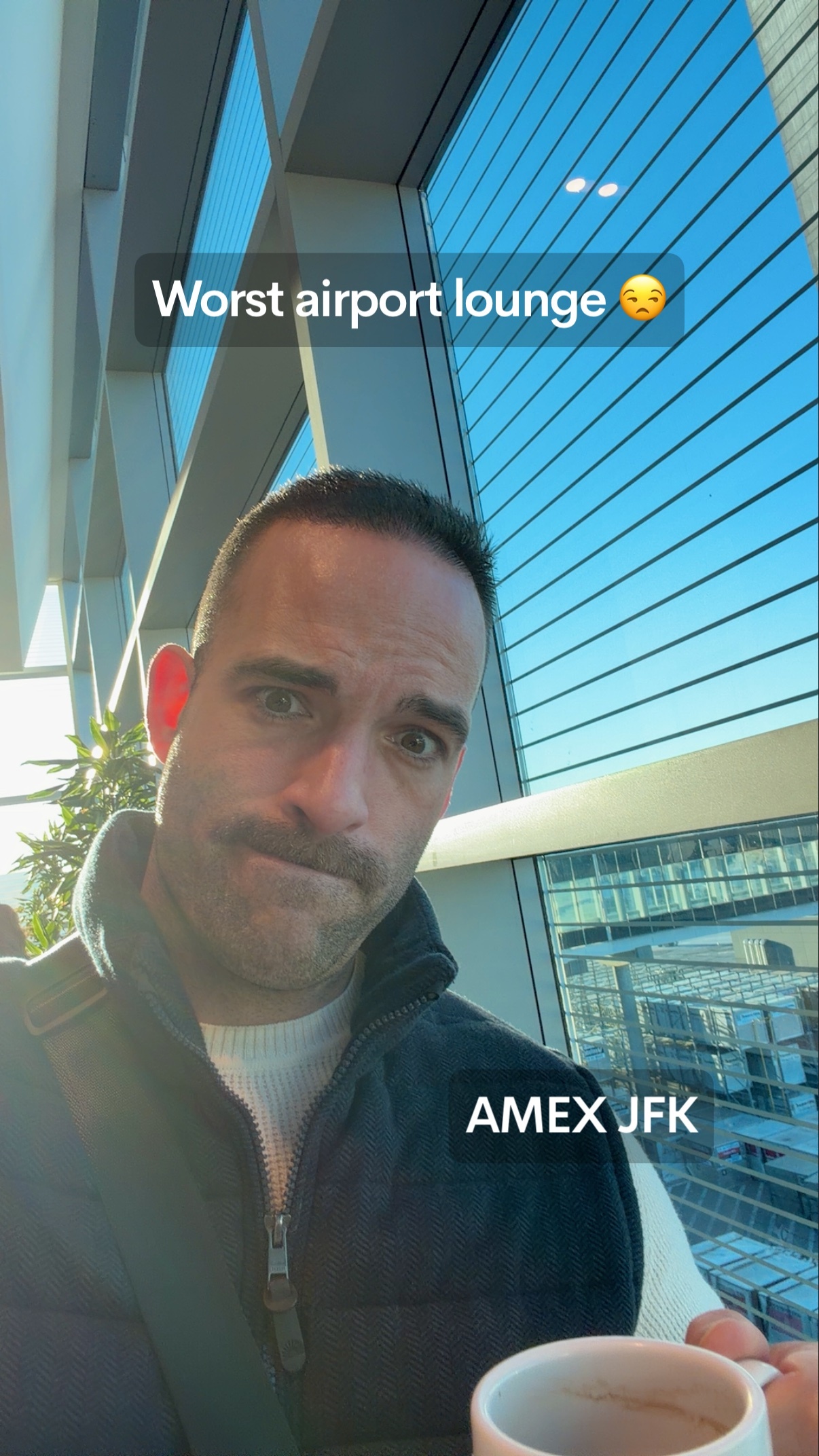 A man with a mustache takes a selfie in an airport lounge, looking displeased. He holds a coffee cup and is seated near a large window with a view of the sky and buildings.