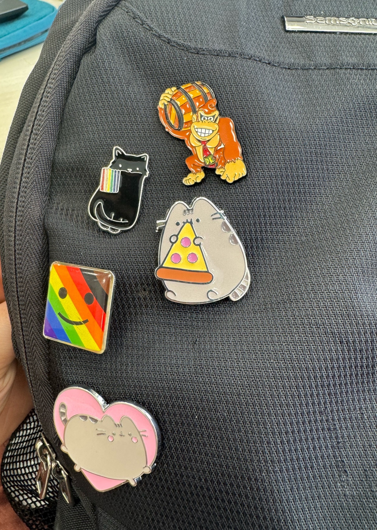 A close-up shot of a black backpack adorned with several colorful enamel pins. The pins feature a black cat with a rainbow stripe on its tail, a rainbow flag square, Donkey Kong holding a barrel, a Pusheen cat eating pizza, and another Pusheen cat in