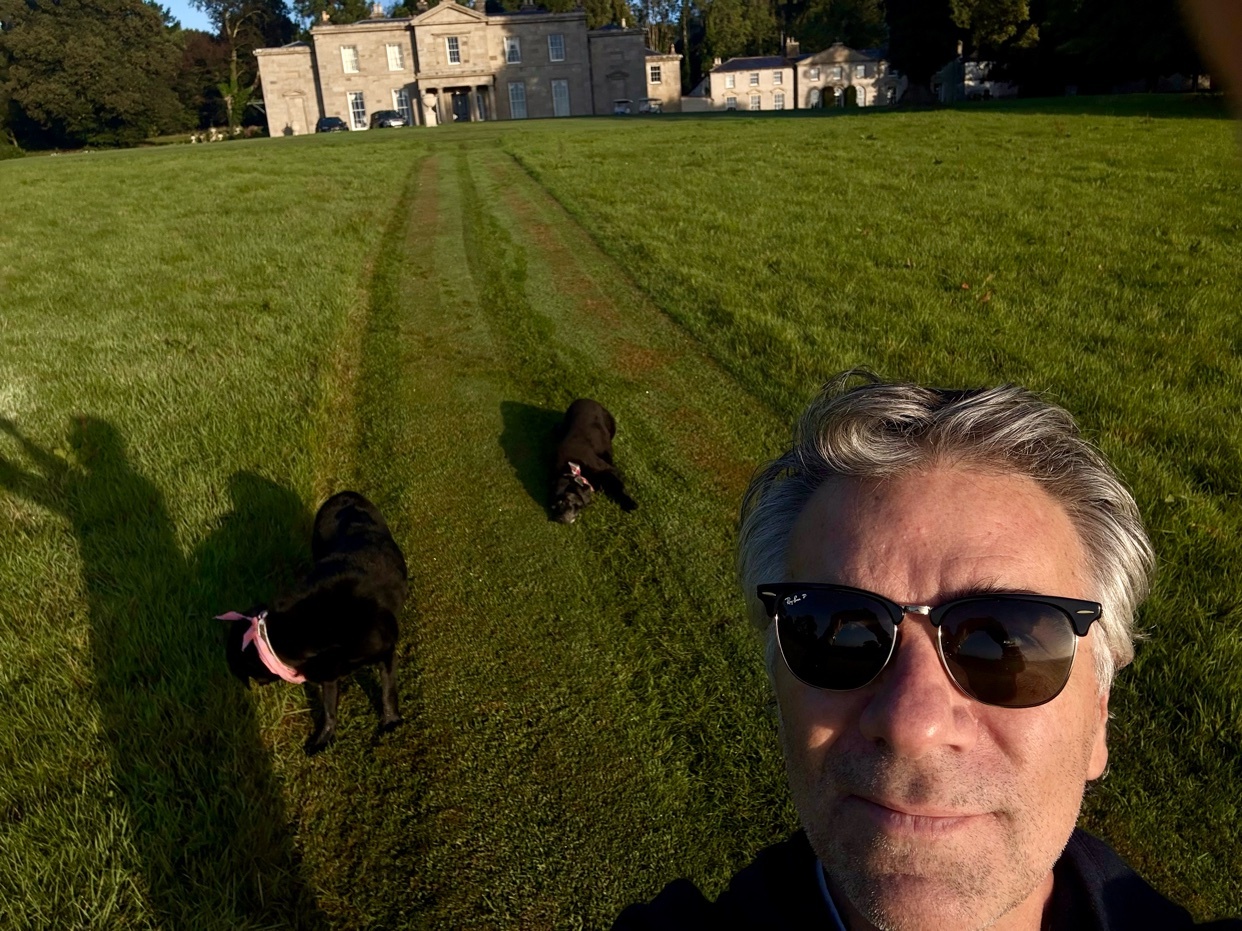 A man takes a selfie in front of a large estate with two black dogs in the grassy yard. The man is wearing sunglasses and the sun casts long shadows across the lawn.