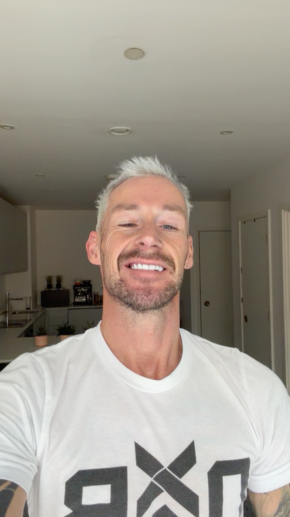 A man with short, grey hair and a bright smile poses for a selfie in what appears to be a kitchen. He is wearing a white t-shirt with a graphic logo.