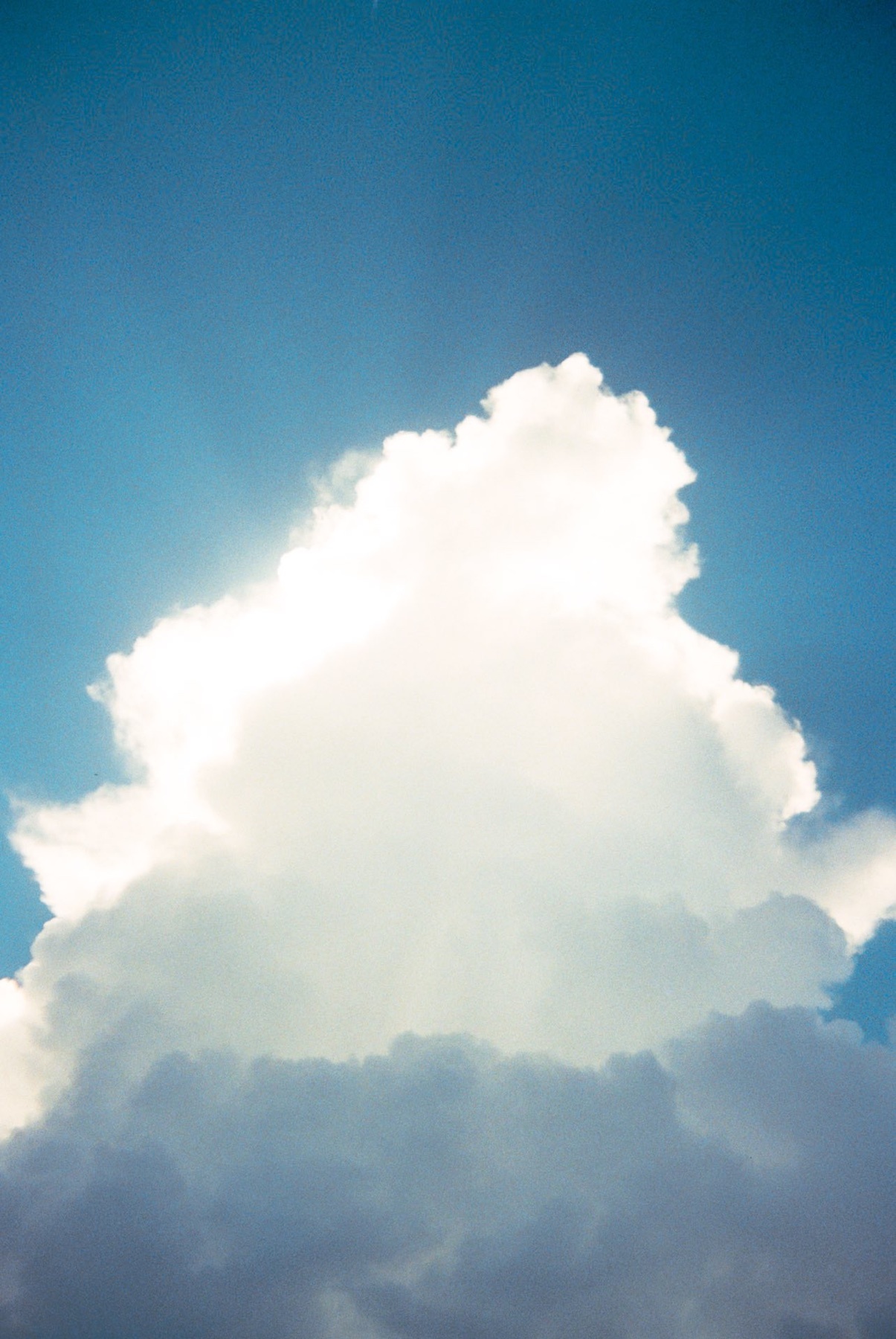 A bright white cumulus cloud is illuminated by the sun, set against a clear blue sky. The sunlight peeks from behind the cloud, creating a radiant glow.