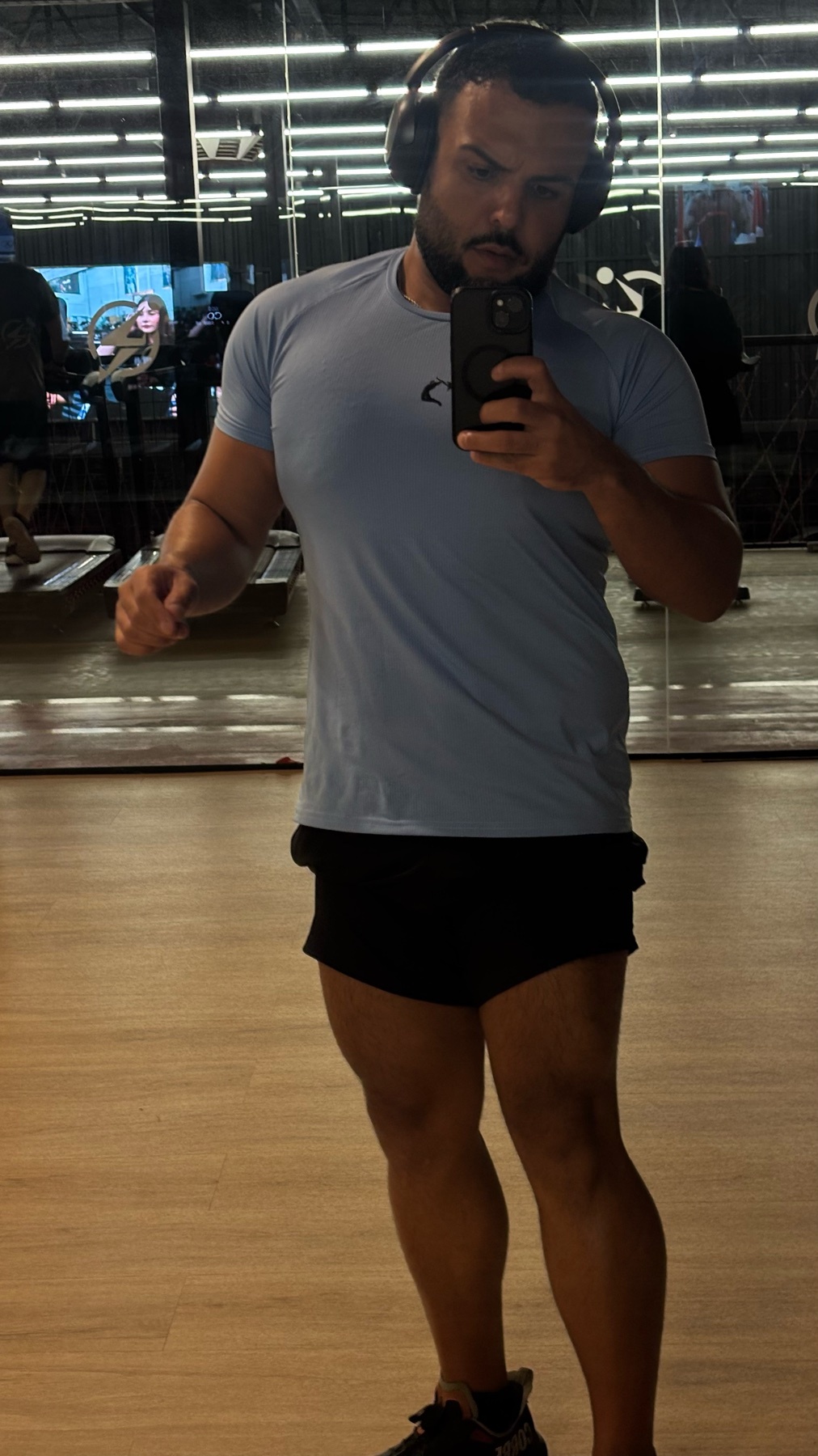 A man in a light blue t-shirt and black shorts takes a selfie in a gym. He is wearing headphones and appears to be working out.