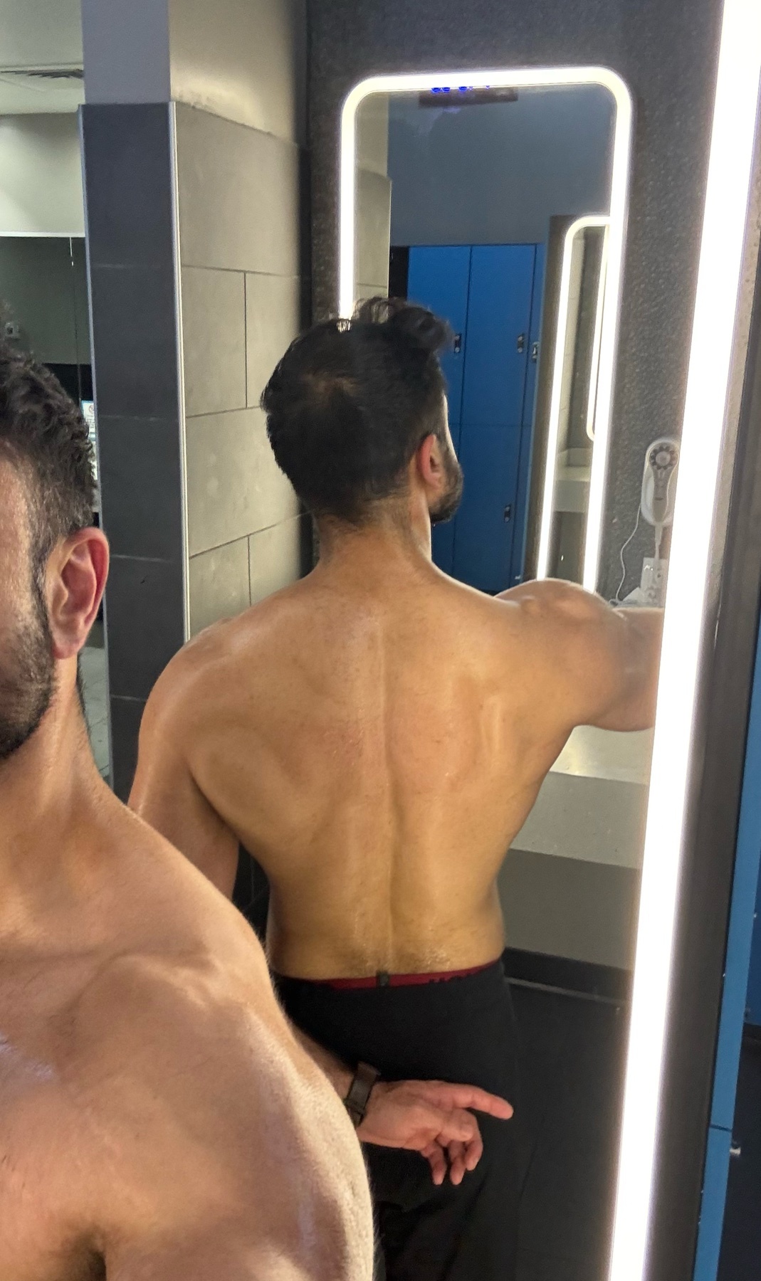 A man is seen from behind, flexing his back muscles in a gym locker room. The lighting highlights his sweaty physique as he poses in front of a mirror.