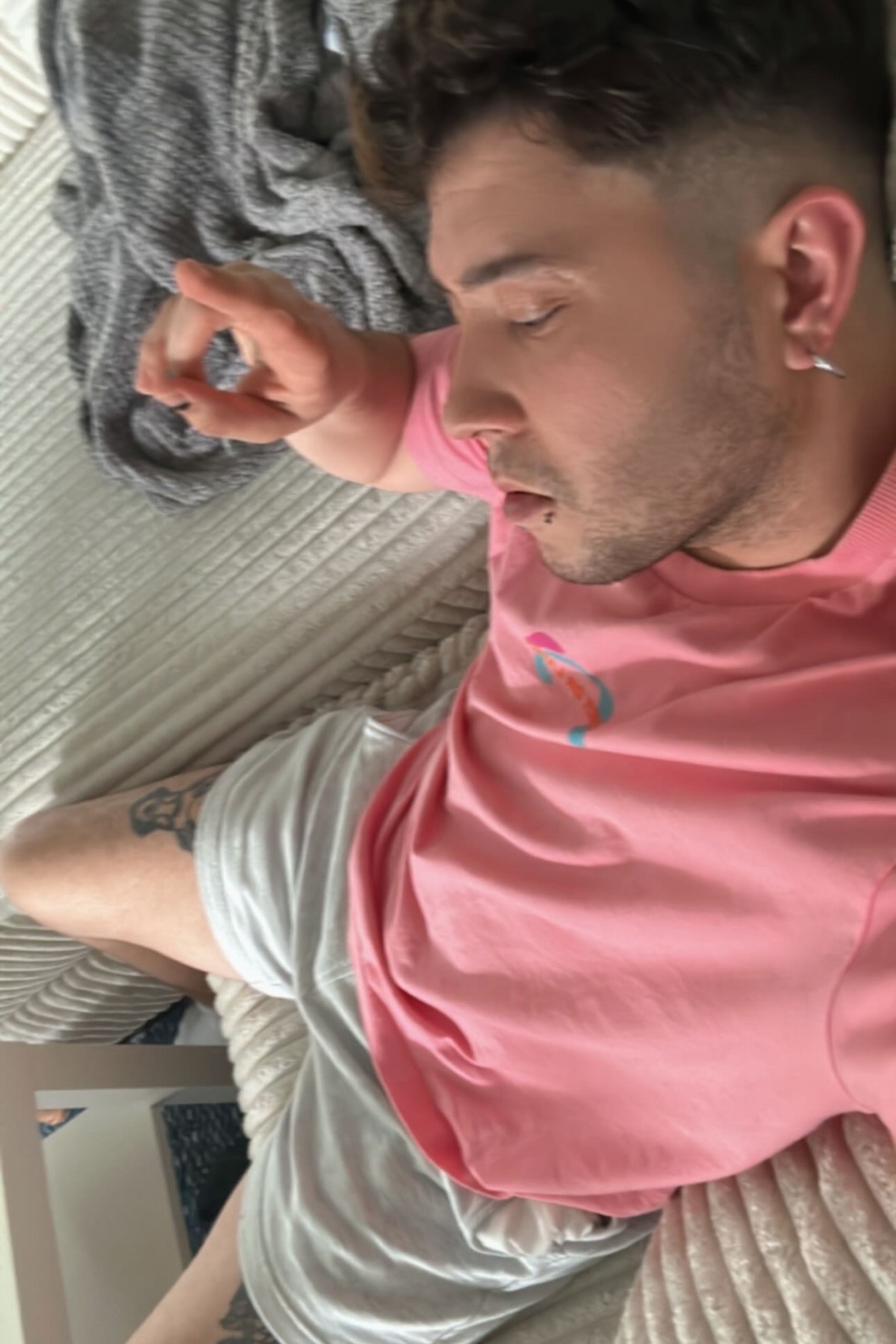 A man is sleeping on a bed, wearing a pink t-shirt and grey shorts. He has short hair and a tattoo on his arm.