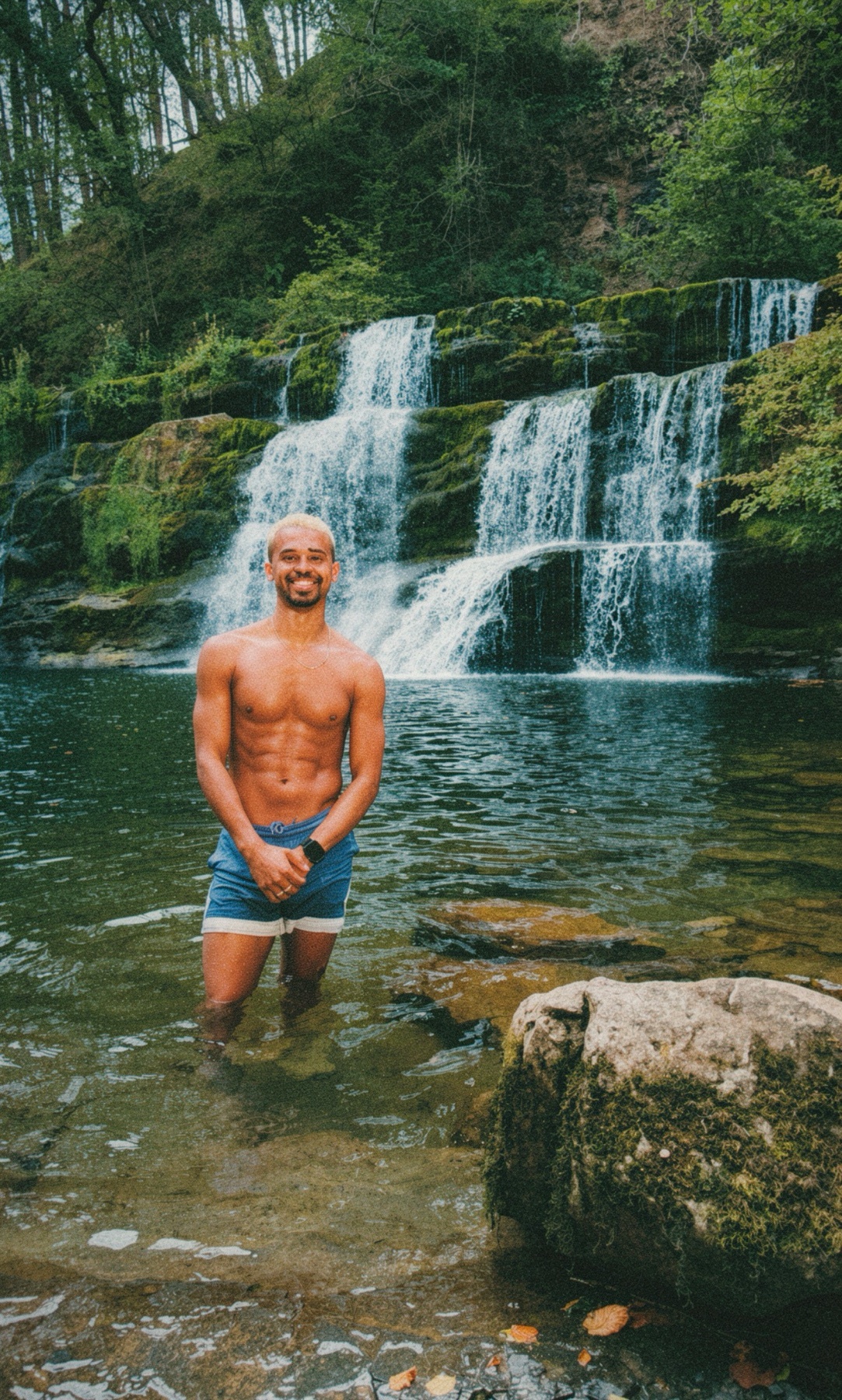 A shirtless man with muscular abs and a beard poses in the water in front of a cascading waterfall surrounded by lush greenery.