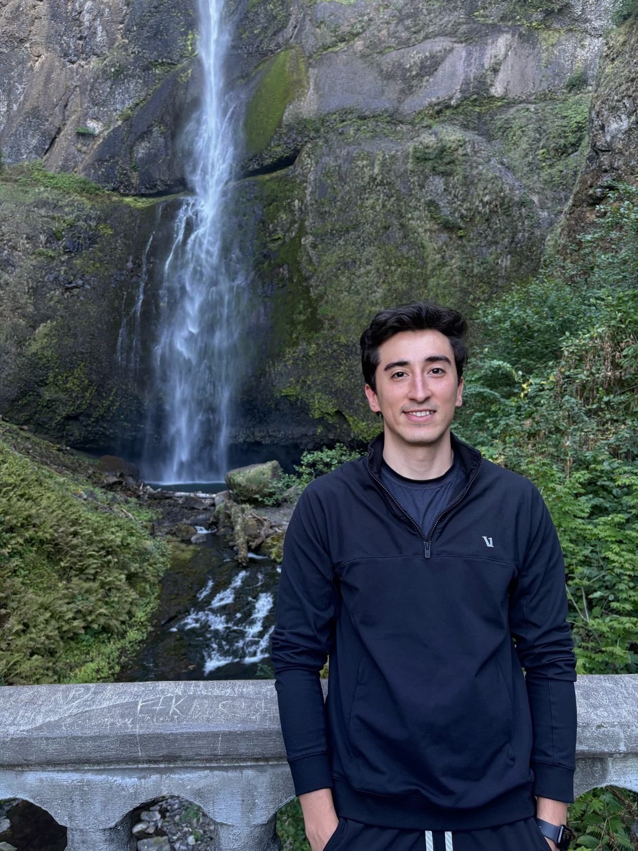 A man poses for a photo in front of a tall waterfall cascading down a rocky cliff face. Lush green foliage surrounds the waterfall and the man stands on a stone bridge.