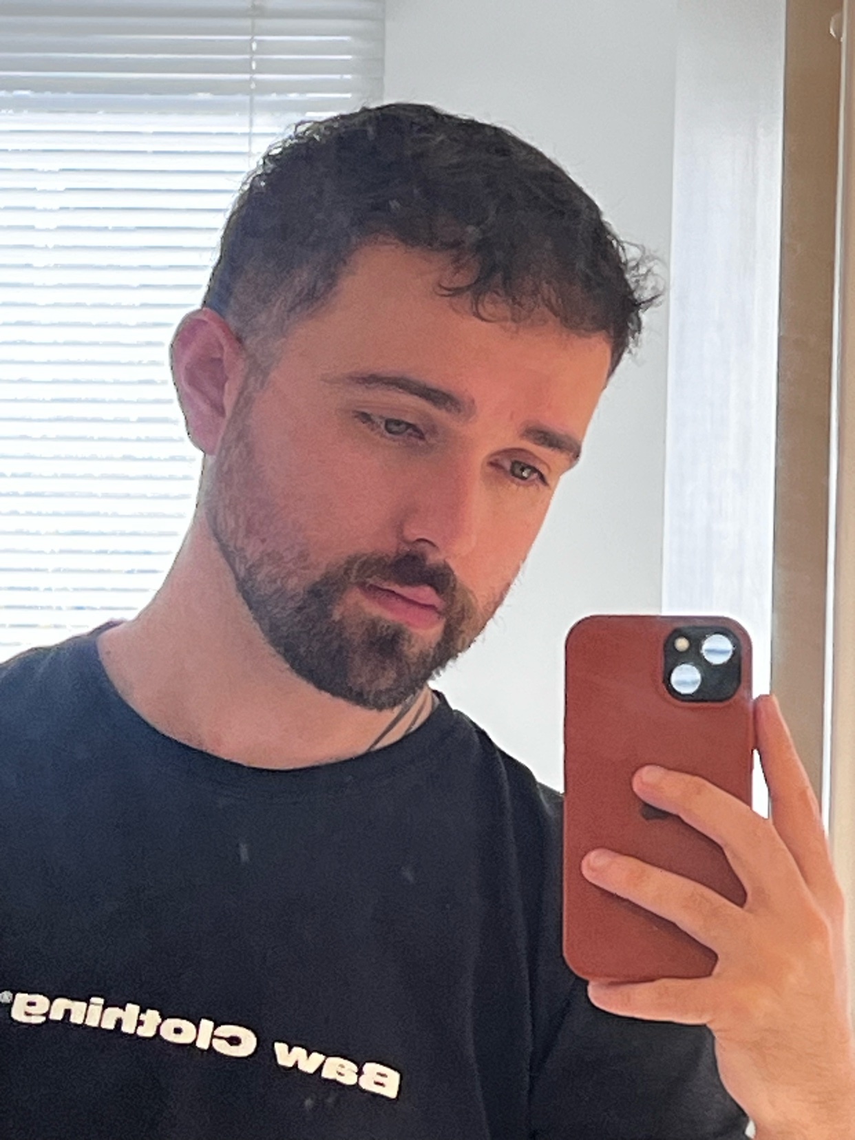 A man with short, dark, curly hair and a beard takes a selfie in a black t-shirt. He is holding a phone in his right hand.