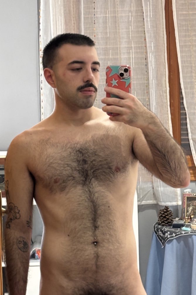 A shirtless man takes a mirror selfie. He has dark hair, a mustache, and a tattoo on his arm.