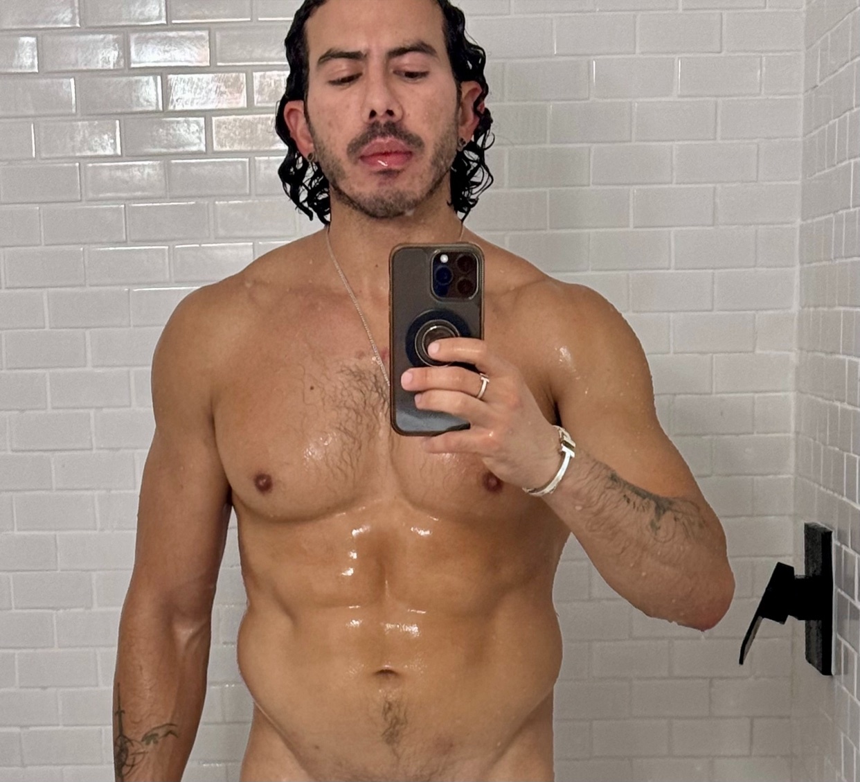 A man with wet, dark, curly hair and stubble takes a selfie in a shower. His muscular, wet torso is visible.