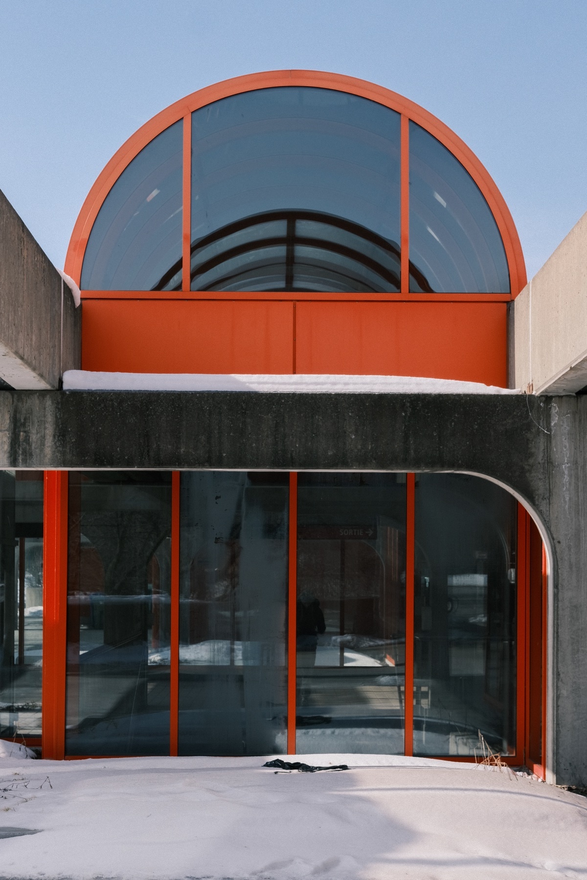 A striking modern architectural design features a large, arched window with an orange frame, set against a concrete facade. The building's exterior is partially covered in snow, suggesting a winter setting.
