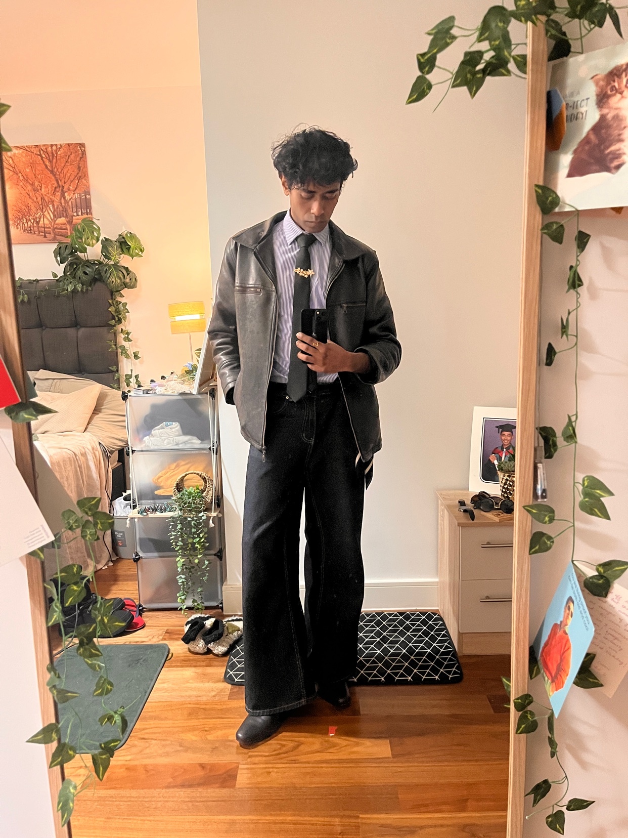 A young man stands in front of a mirror, wearing a leather jacket over a collared shirt and tie, with wide-leg pants and dress shoes. The room is decorated with plants and personal items.