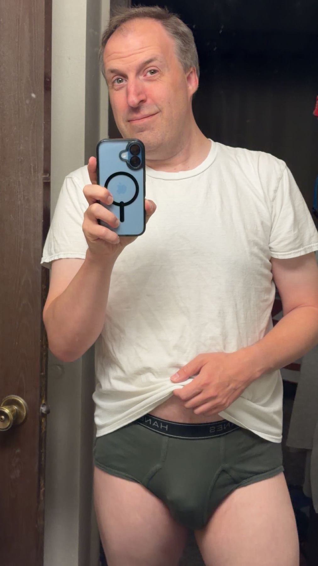 A man in a white t-shirt and green briefs poses for a selfie in a bathroom, holding his phone in his left hand.