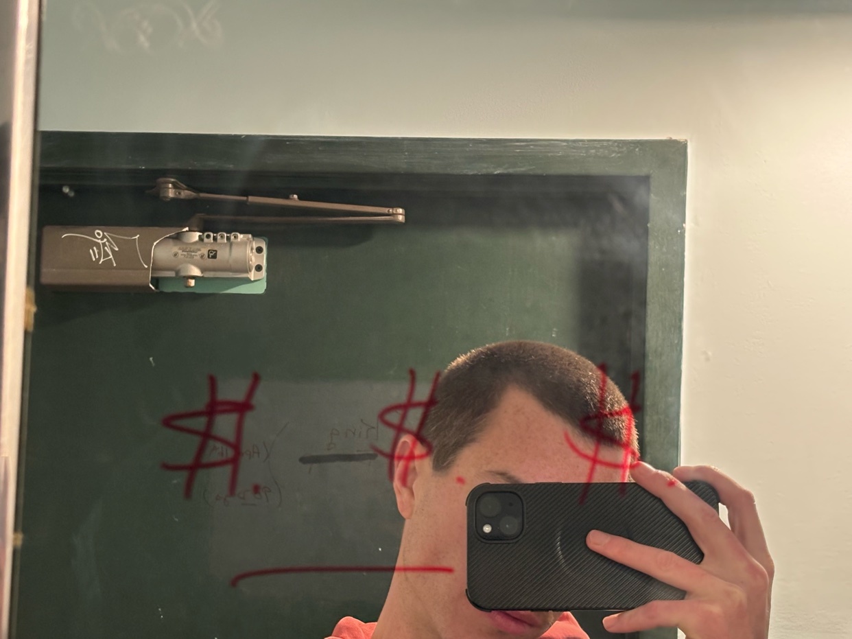 A man takes a selfie in a mirror with graffiti on it. The mirror is in a bathroom.