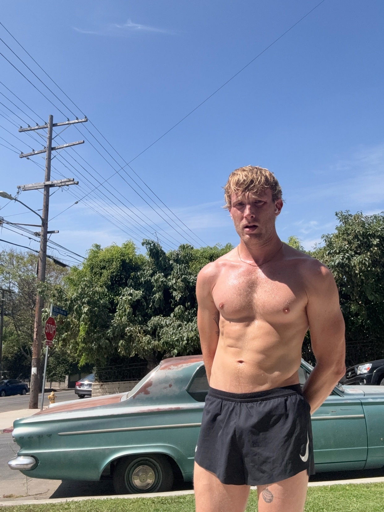 A shirtless man with blonde hair and muscular physique stands outside in running shorts. He is looking towards the camera with a slight grimace, possibly from exertion or the bright sun. Behind him, an old teal car is parked on a residential street u