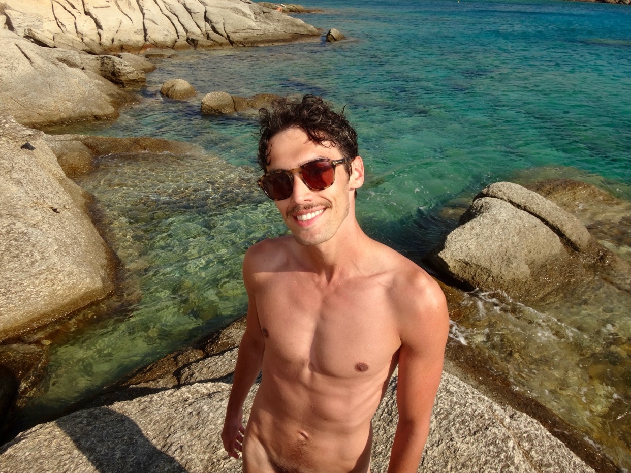 A man poses shirtless on a rocky beach, smiling at the camera. The turquoise water and rocks create a scenic backdrop.