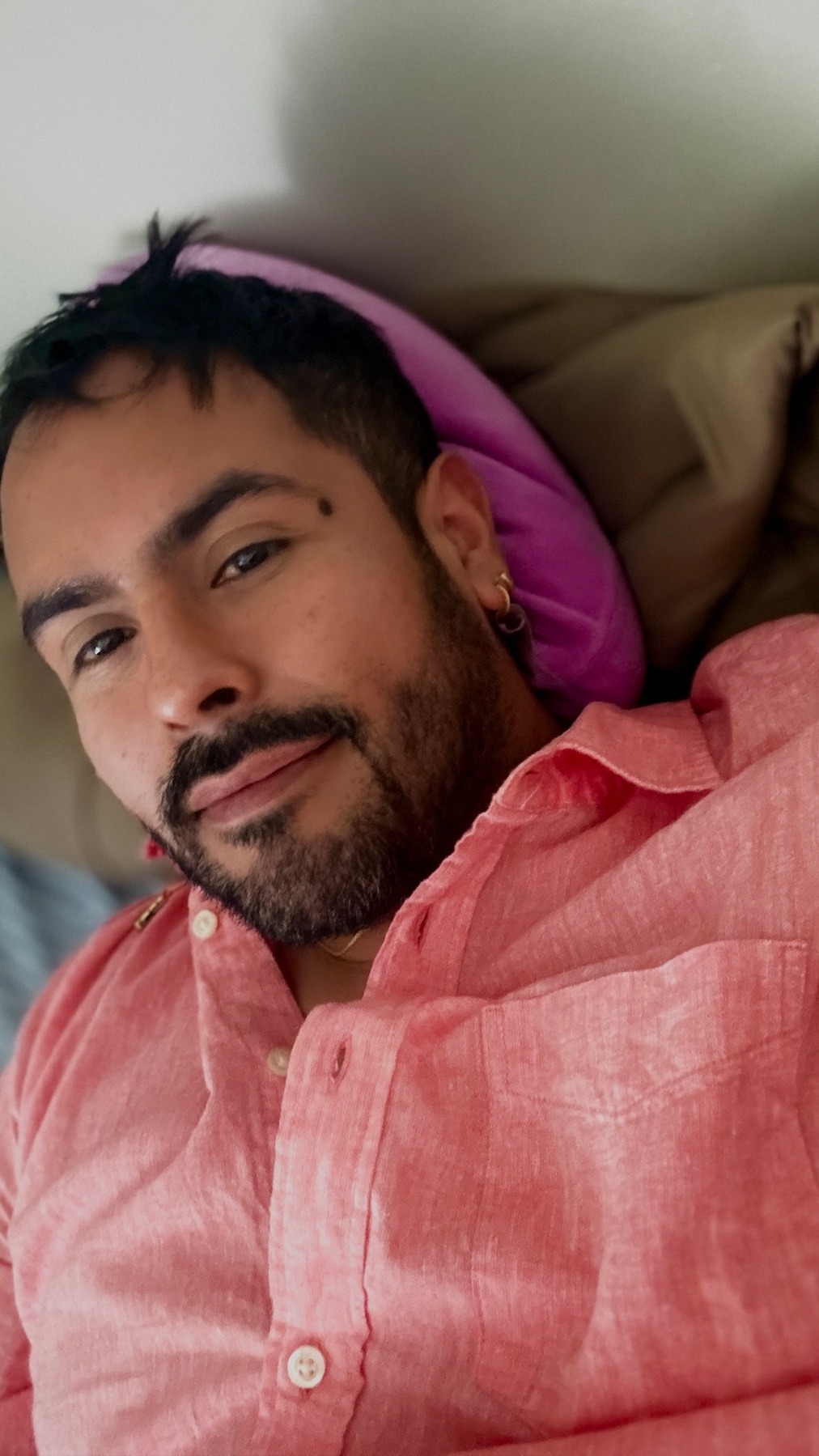 A man poses for a selfie, wearing a pink shirt. He has dark hair and a beard.