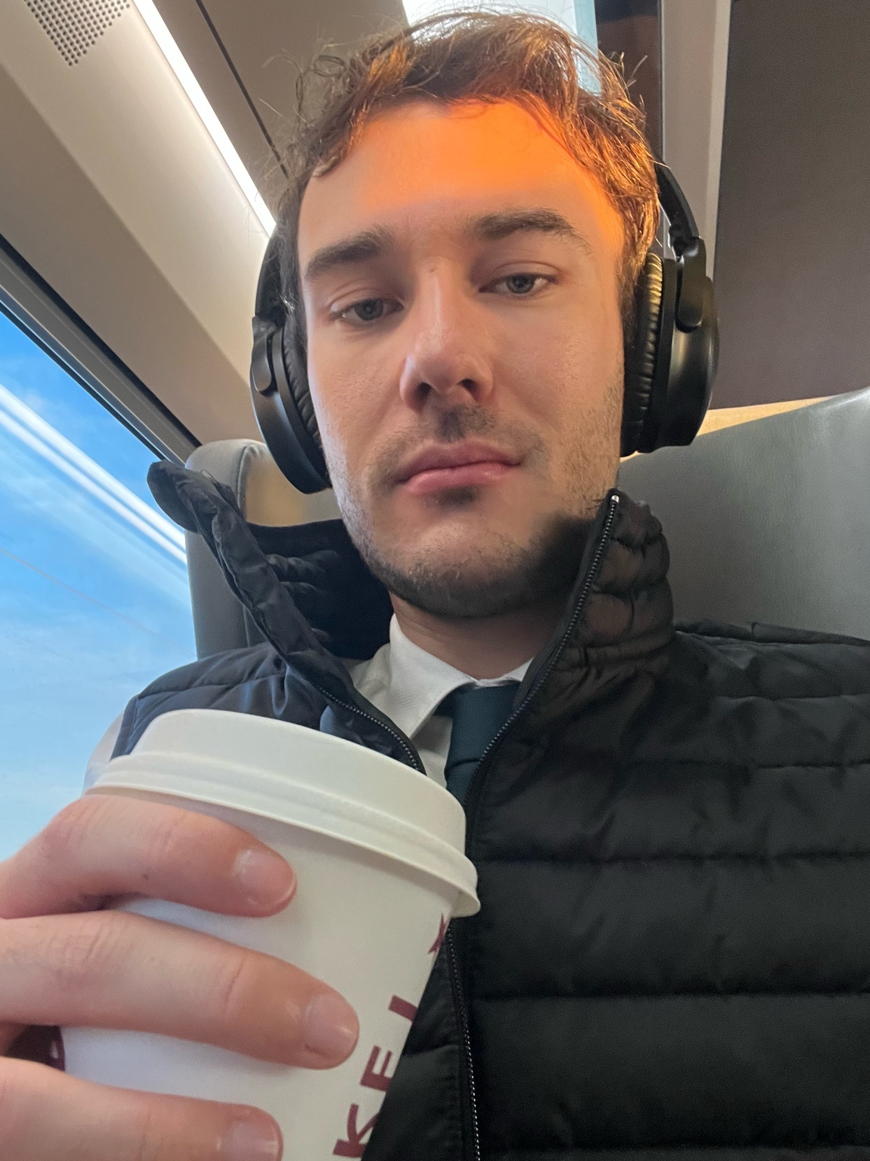 A man takes a selfie while holding a coffee cup and wearing headphones. He is dressed in business casual attire, suggesting he is on his commute.