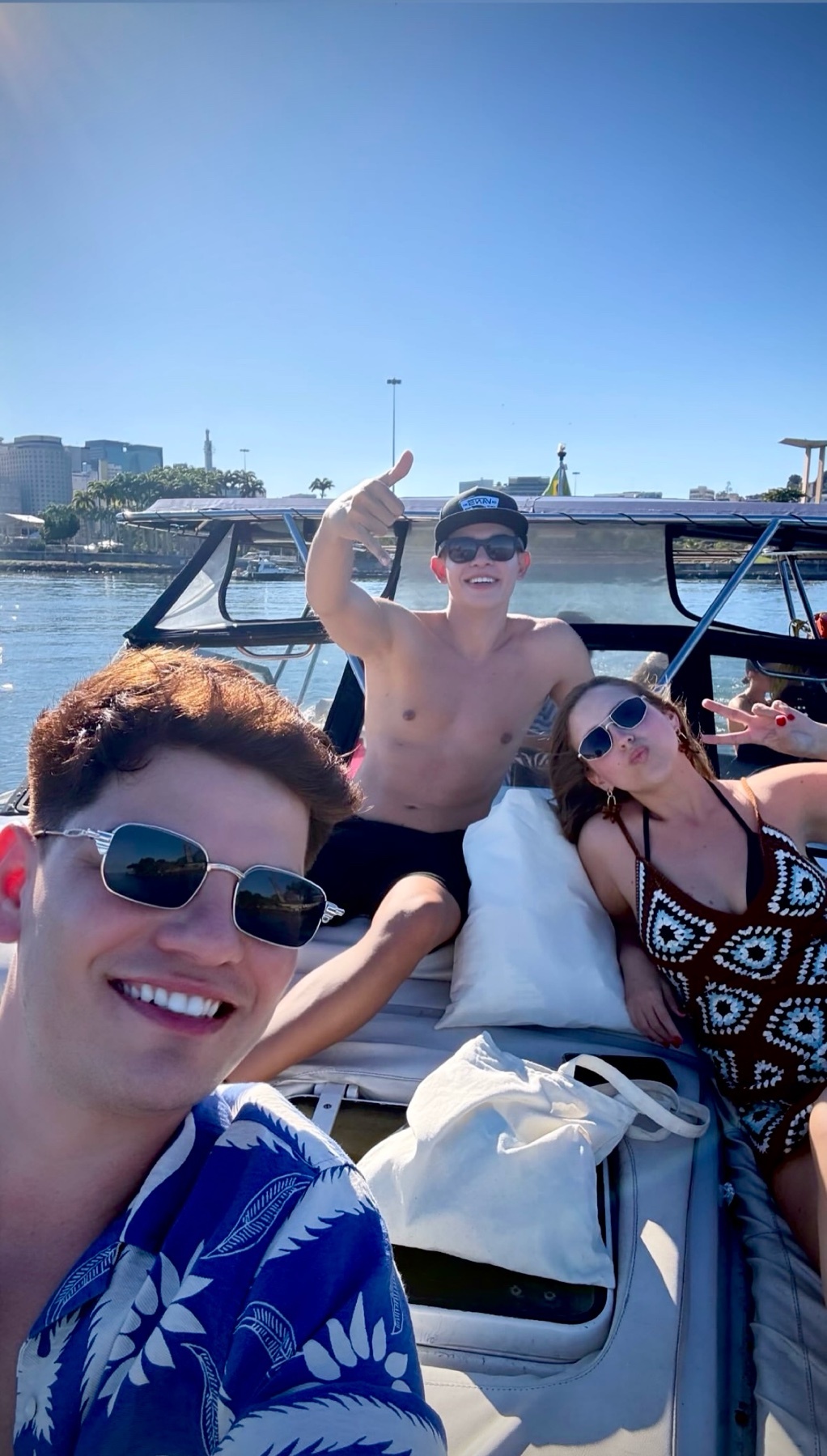 Three friends are enjoying a sunny day on a boat trip. One person in the foreground smiles at the camera, wearing a blue floral shirt and sunglasses. In the background, two others pose, one giving a thumbs up and the other making a peace sign.