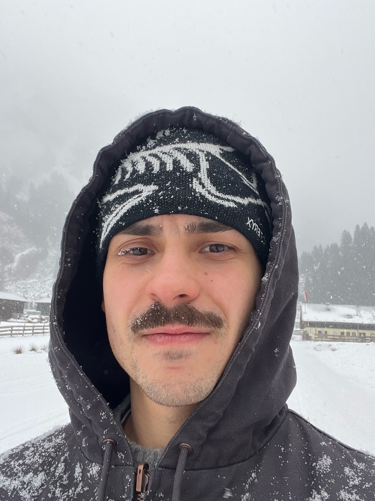 A man in a hoodie poses for a selfie in the snow. The man has a mustache and is wearing a beanie.