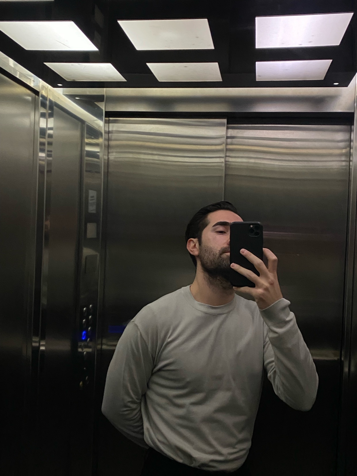 A man takes a selfie in an elevator, looking into the camera with a beard and dark hair.