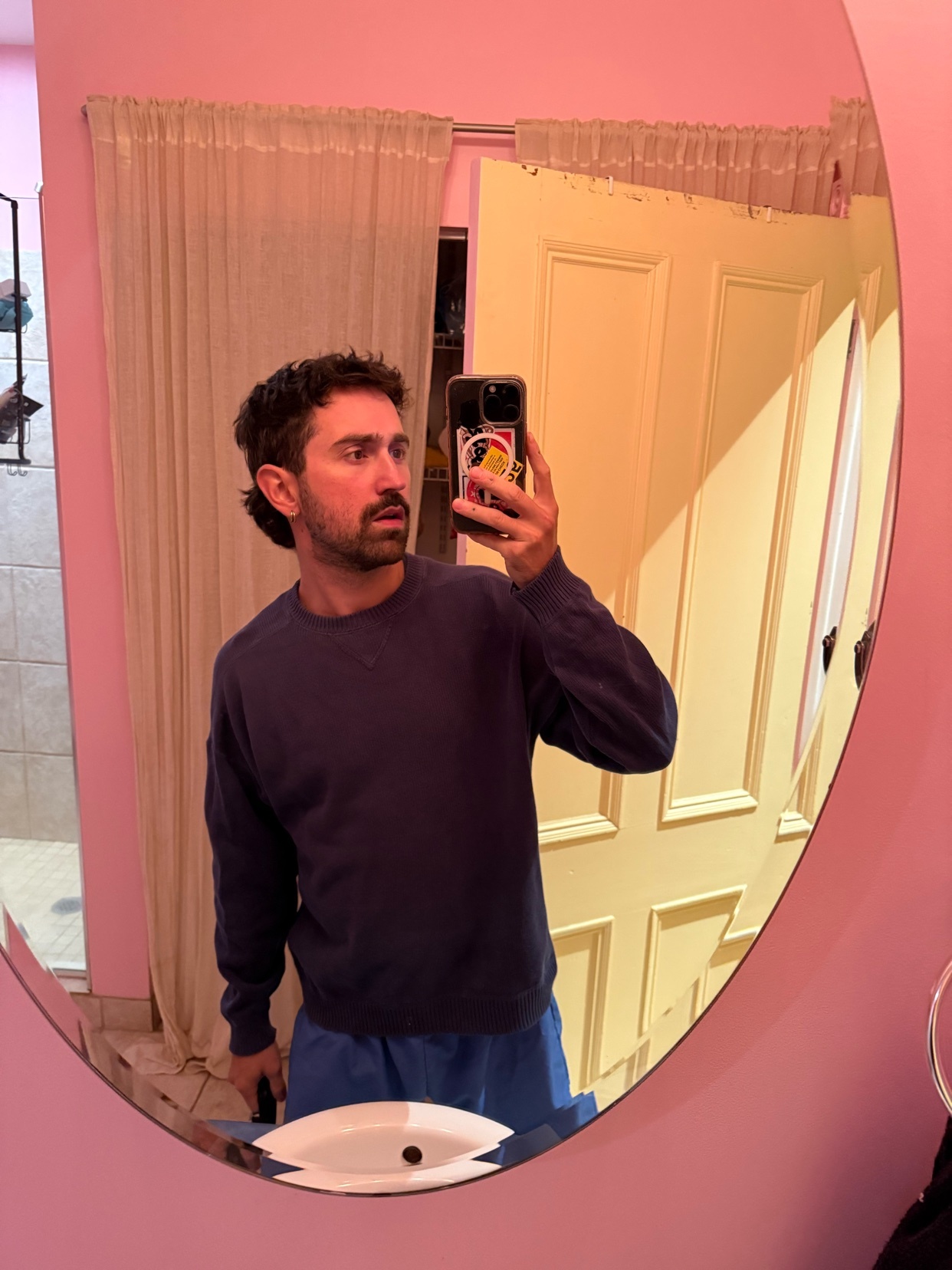 A man with dark, curly hair and stubble takes a mirror selfie in a pink-tiled bathroom. He is wearing a navy blue sweatshirt and blue shorts.