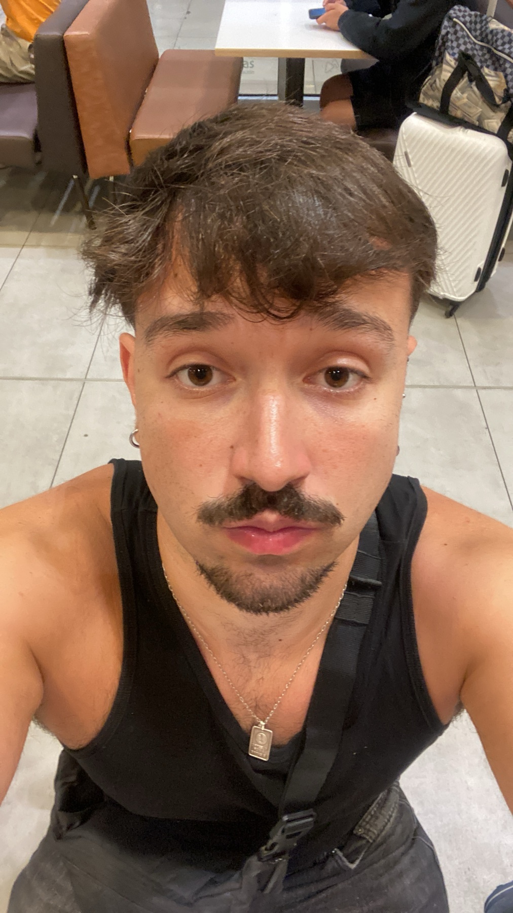 A close-up selfie of a man with a mustache and stubble, wearing a black tank top and a necklace. He appears to be in an airport with luggage in the background.