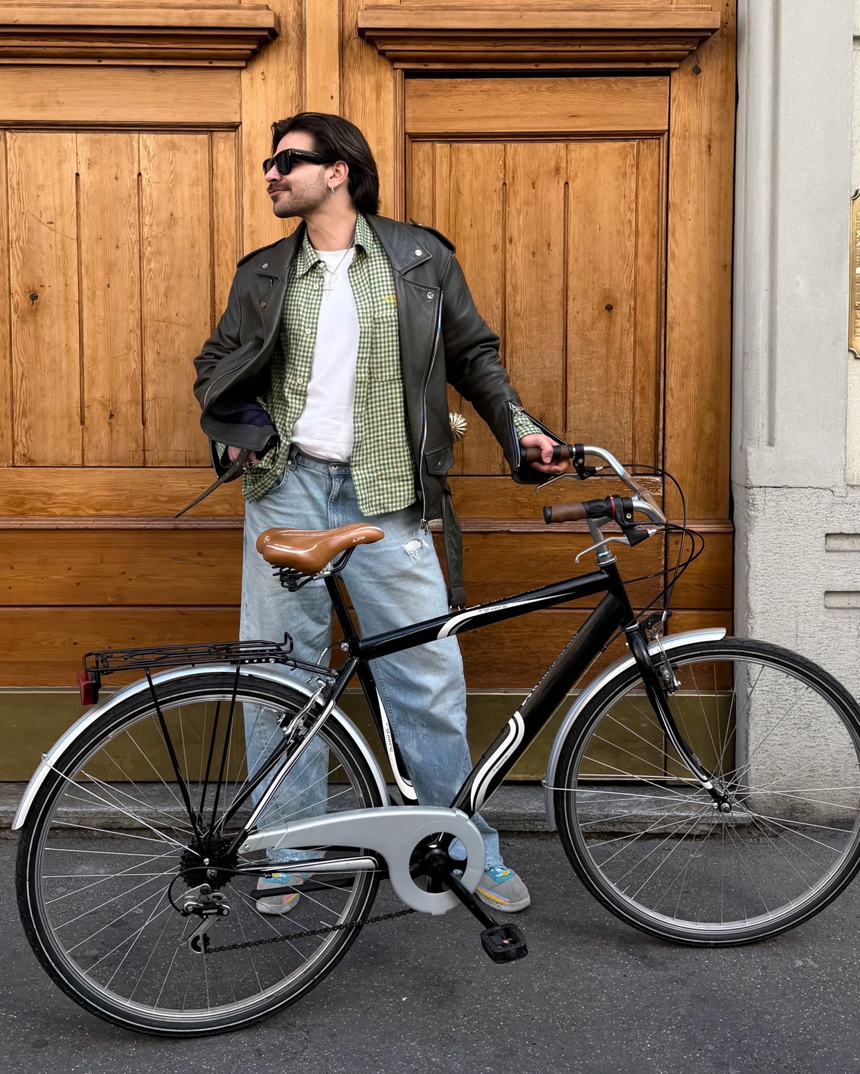 A man in a leather jacket and sunglasses poses with his bicycle in front of a wooden door.