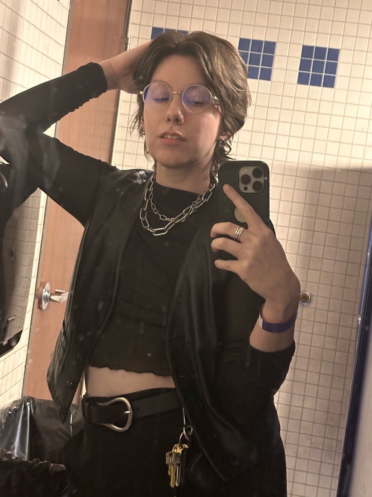 A person with short, dark hair and glasses takes a selfie in a bathroom. They are wearing a black outfit with layered clothing and silver jewelry.