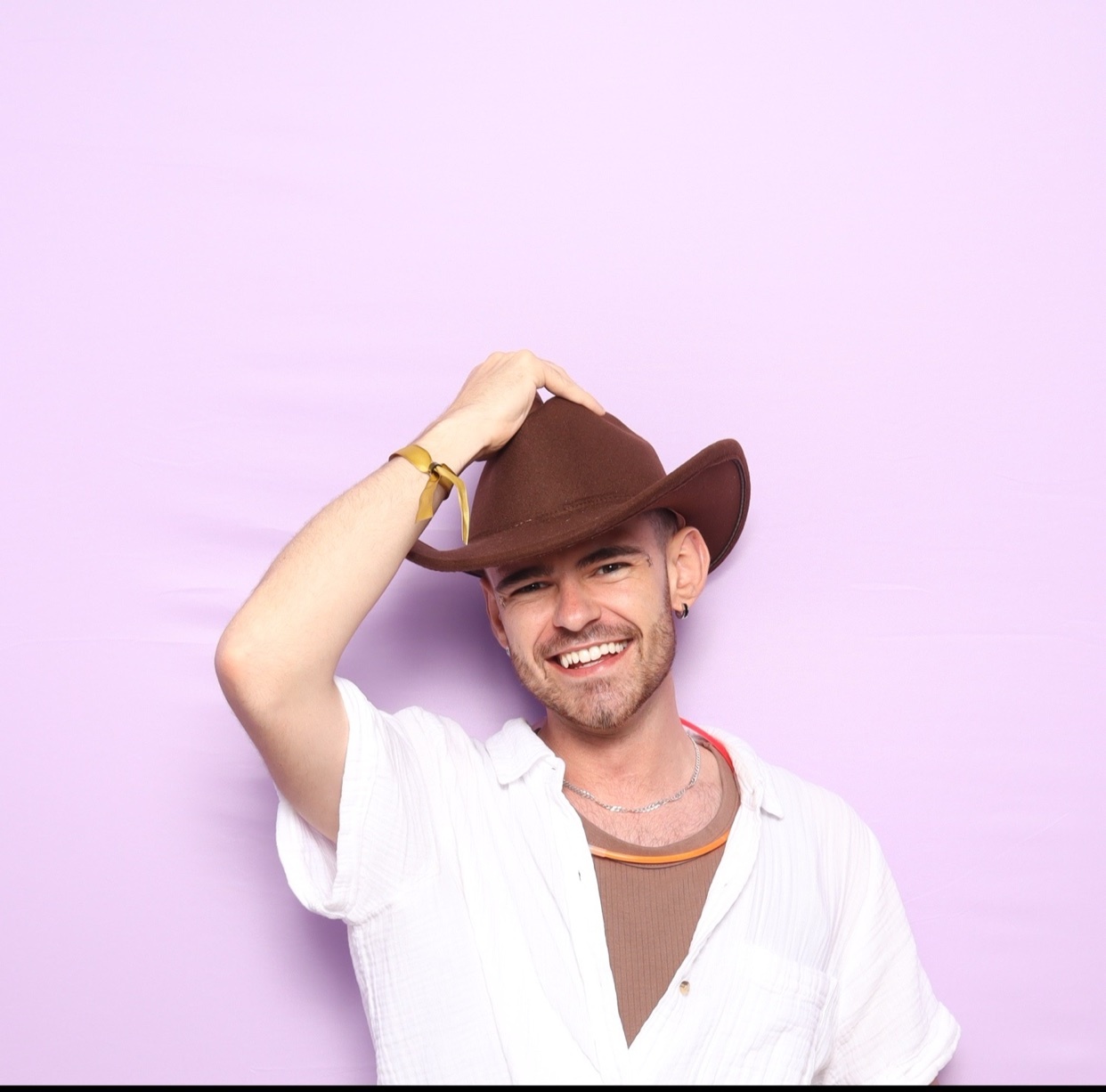 A man in a cowboy hat smiles while posing in a photo booth against a lavender background. He is wearing a white button-down shirt over a brown t-shirt and accessorized with a necklace and bracelet.