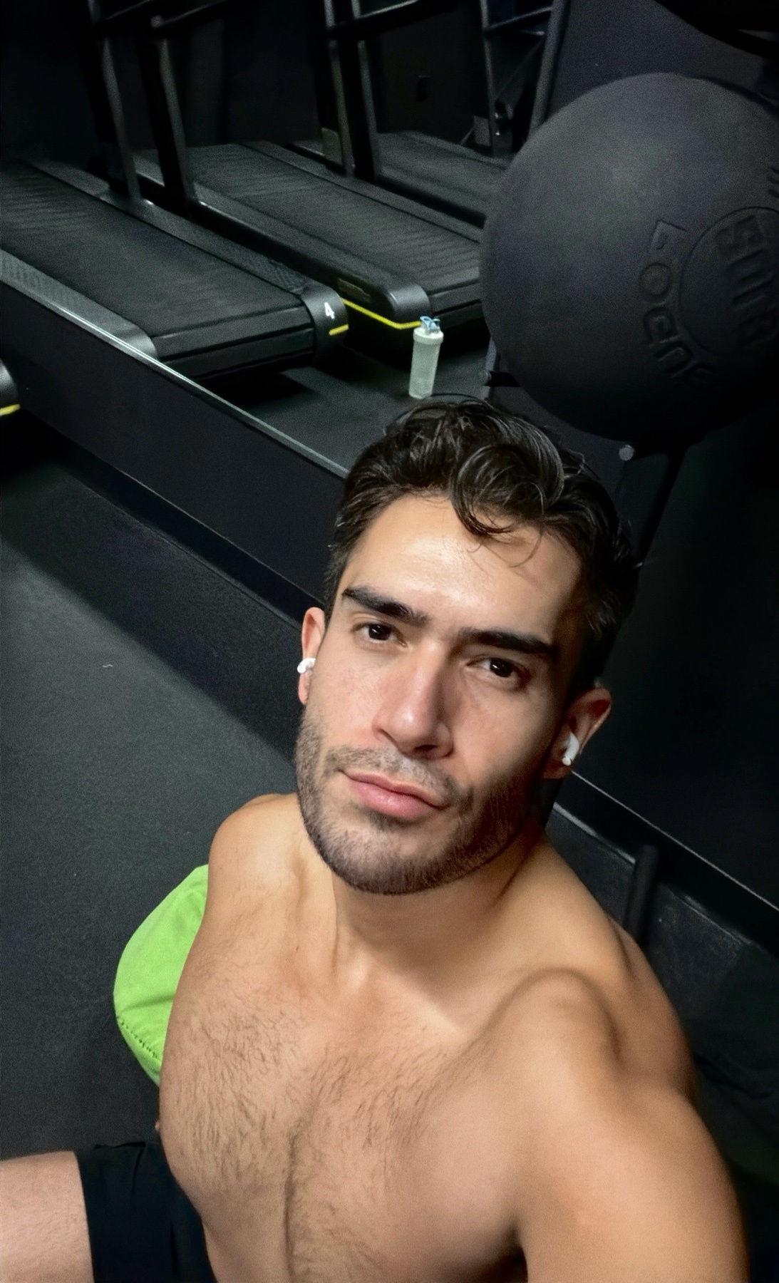 A man takes a selfie at the gym, shirtless and looking at the camera. The background includes treadmills and a medicine ball.