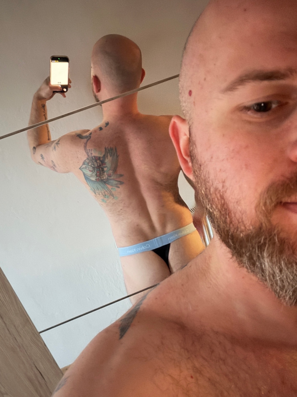 A man with a beard and tattoos takes a mirror selfie while wearing a thong and no shirt.