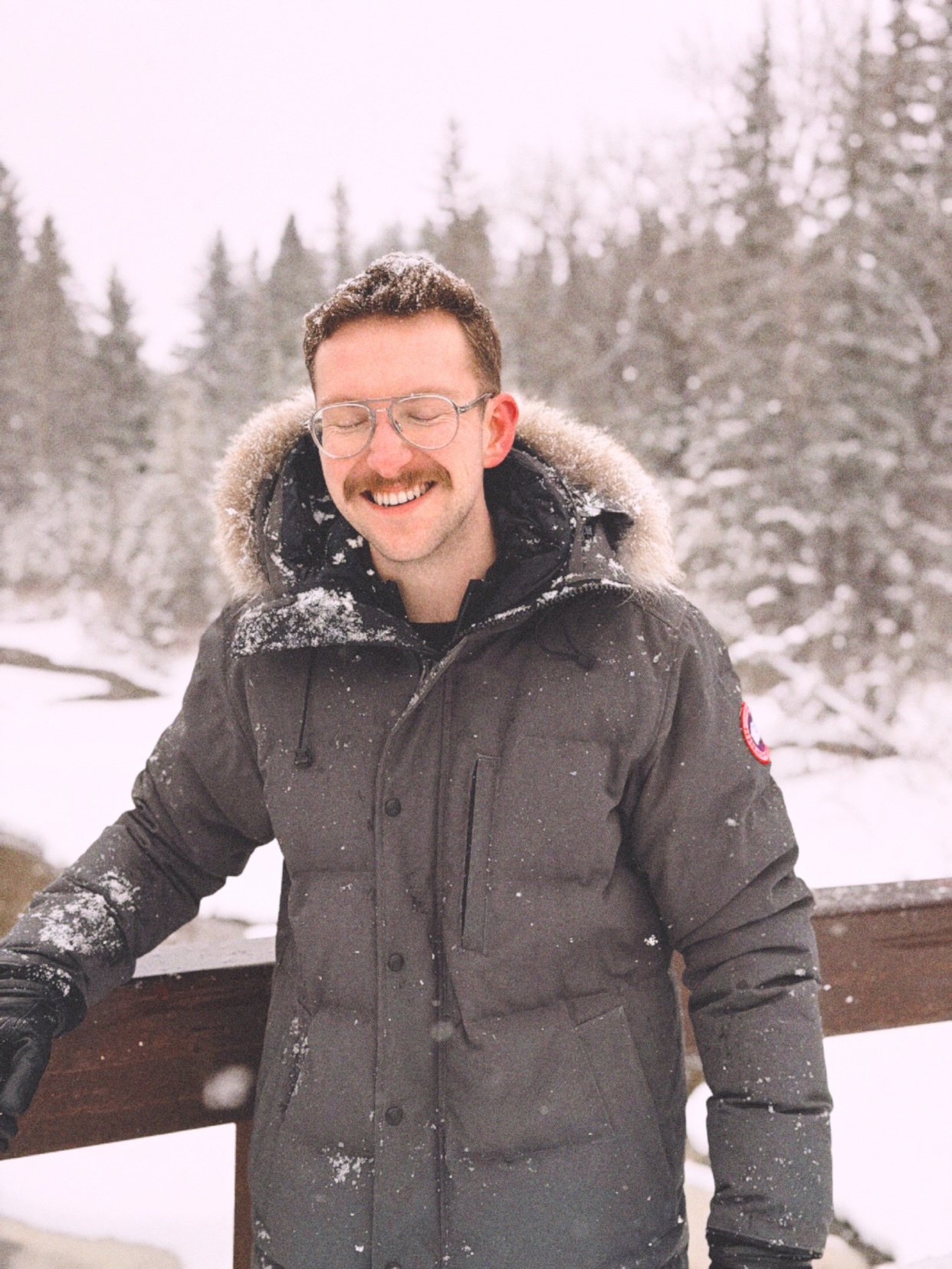 A man with a mustache and glasses smiles while wearing a gray parka with a fur-lined hood in a snowy, wooded area.