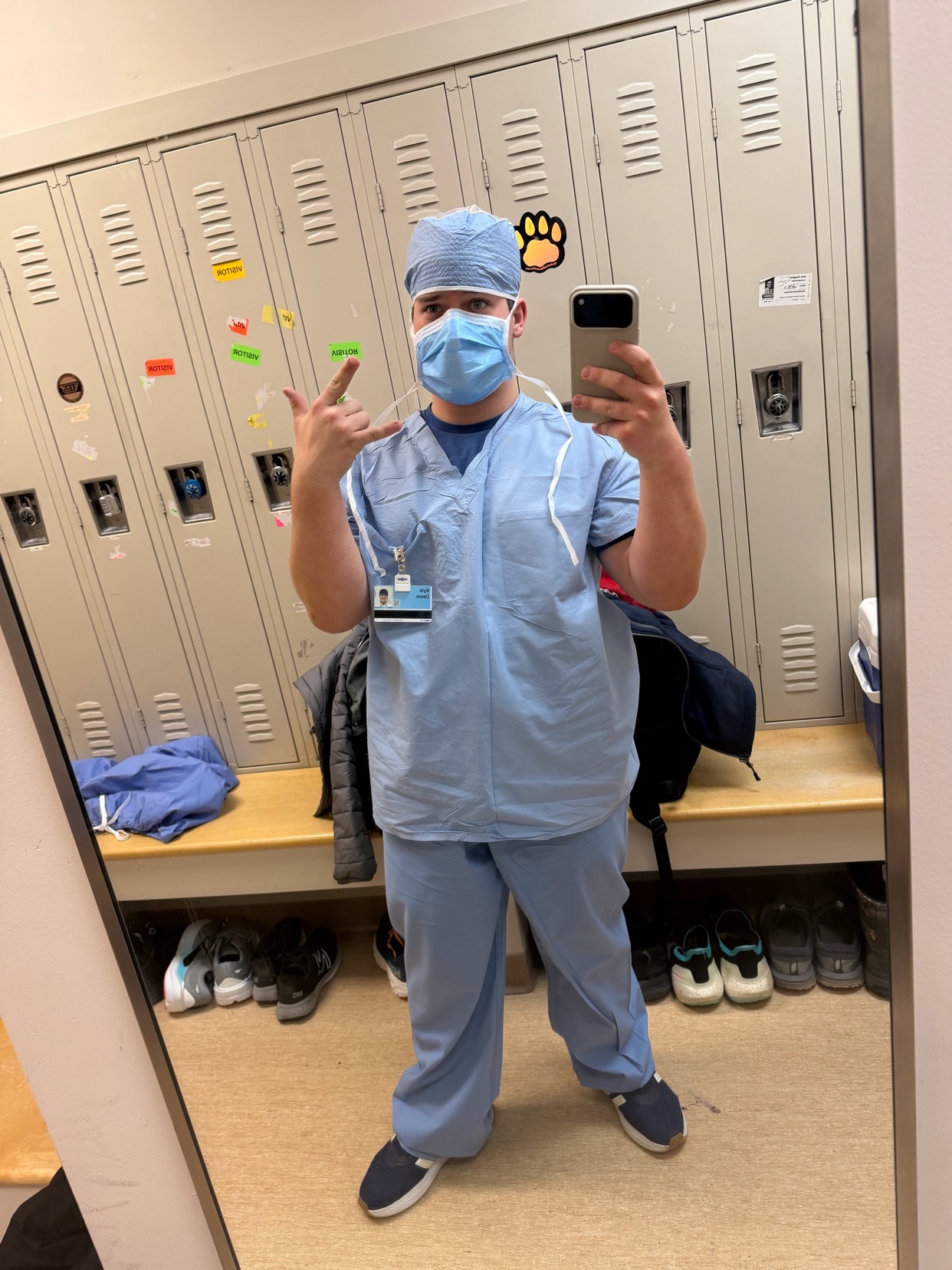 A man in blue scrubs and a scrub cap takes a selfie in a locker room. He is wearing a face mask and has a stethoscope around his neck.