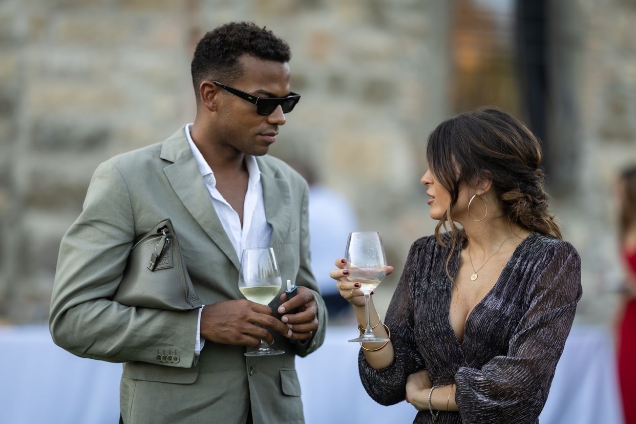 A man and a woman are seen talking while holding glasses of wine. They appear to be at a wedding, with an outdoor setting in the background.
