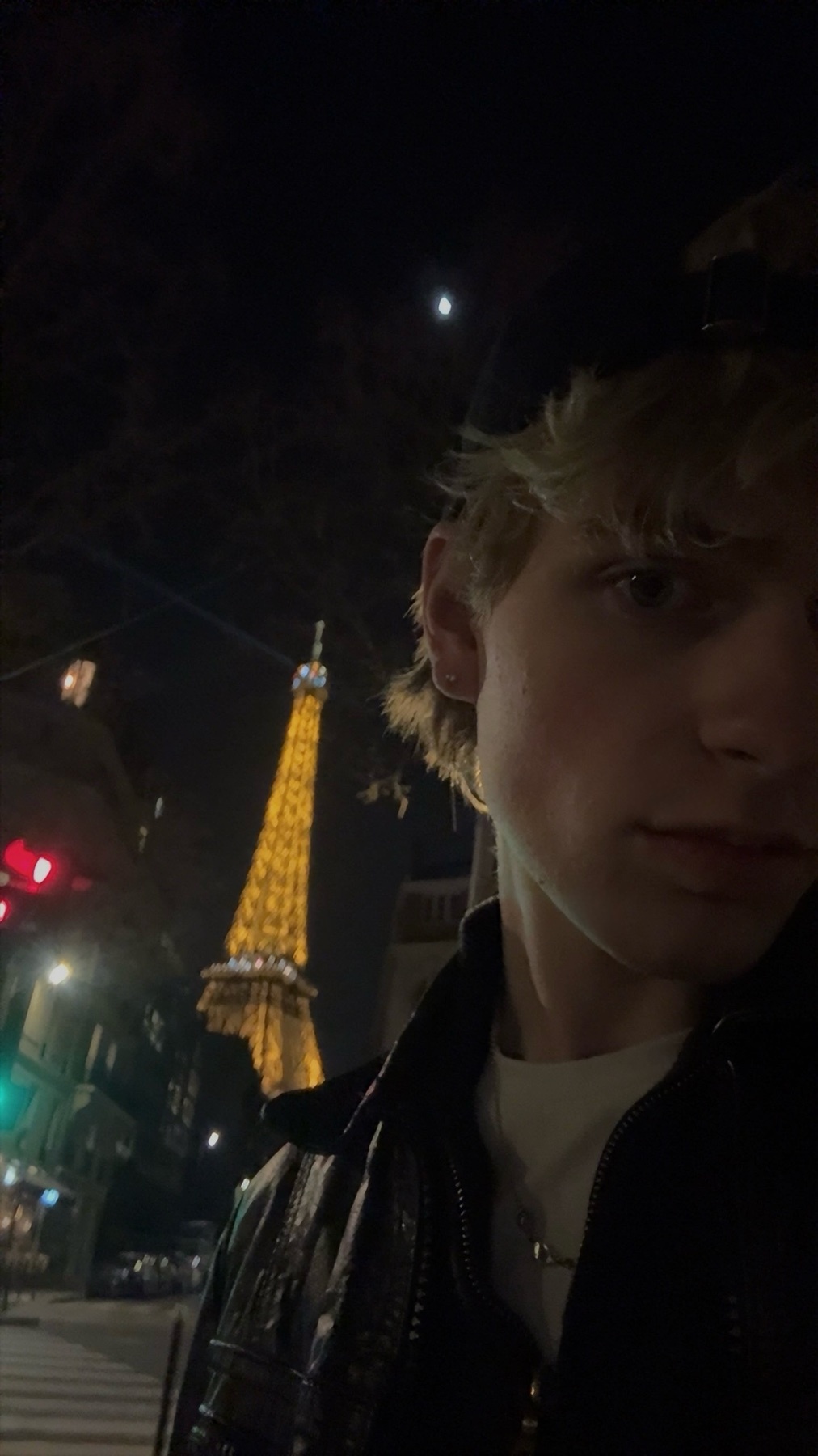 A young man with blond hair takes a selfie at night in Paris, with the Eiffel Tower illuminated in the background.