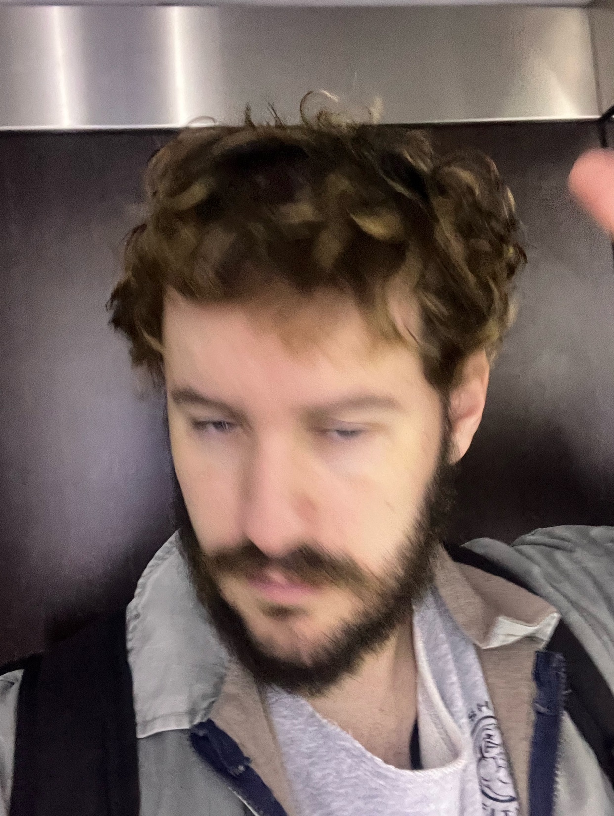 A man with curly hair and a beard takes a selfie. The photo is taken indoors.
