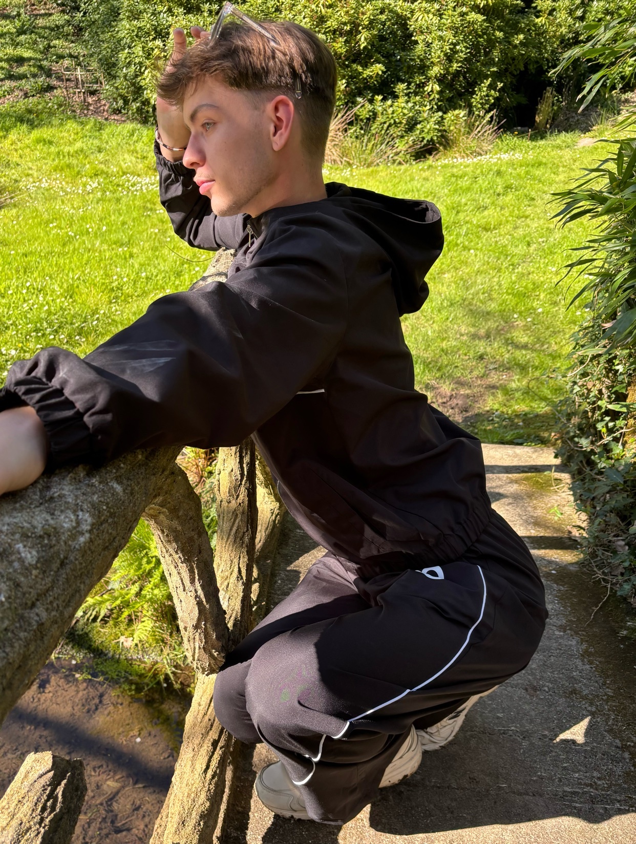 A young man in a black tracksuit poses outdoors in a park, leaning on a wooden railing with a lush green background.