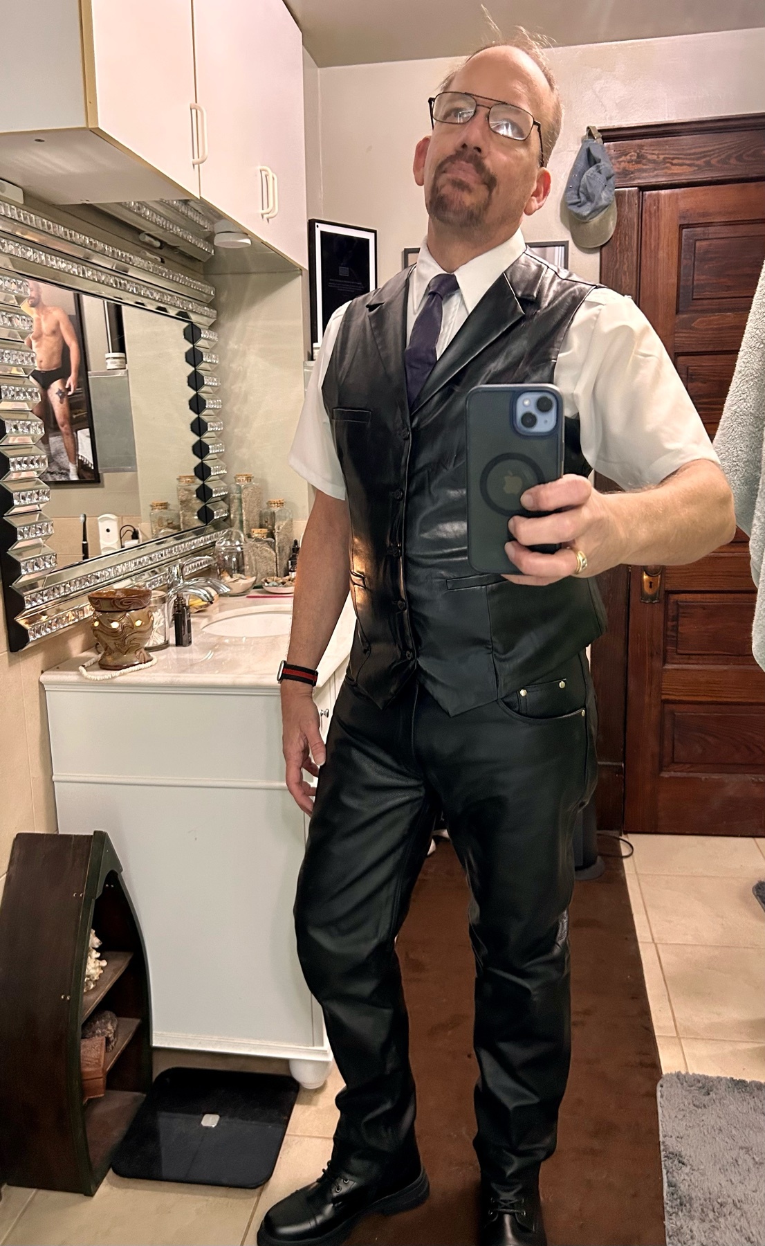 A man in a leather vest, leather pants, a white button-down shirt, and a tie poses for a mirror selfie. He is wearing glasses and black boots.